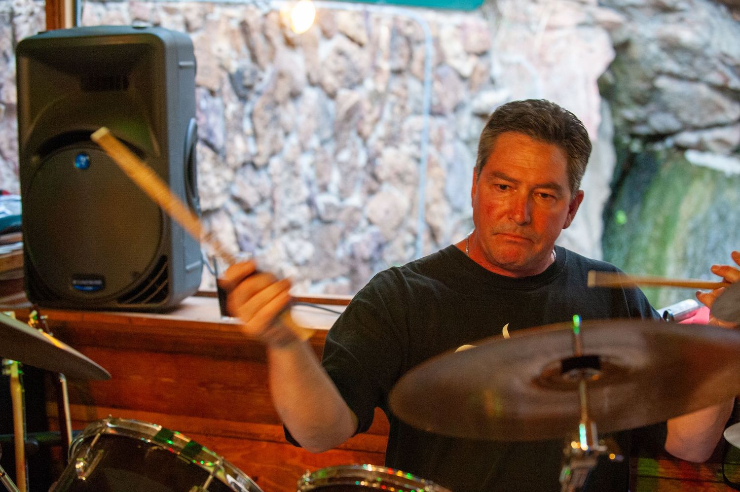 A man playing drums with sticks at an outdoor venue, with a speaker and stone wall in the background.
