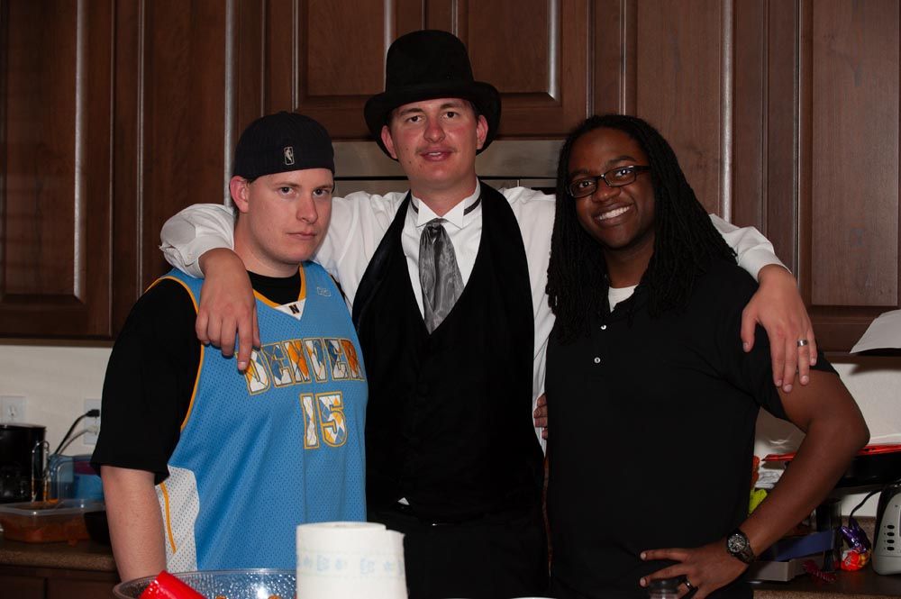 Three people pose together at a party, with one wearing a top hat and formal vest.