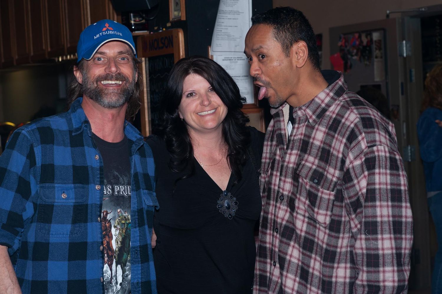 Three people pose together in casual attire including plaid shirts at an indoor venue.