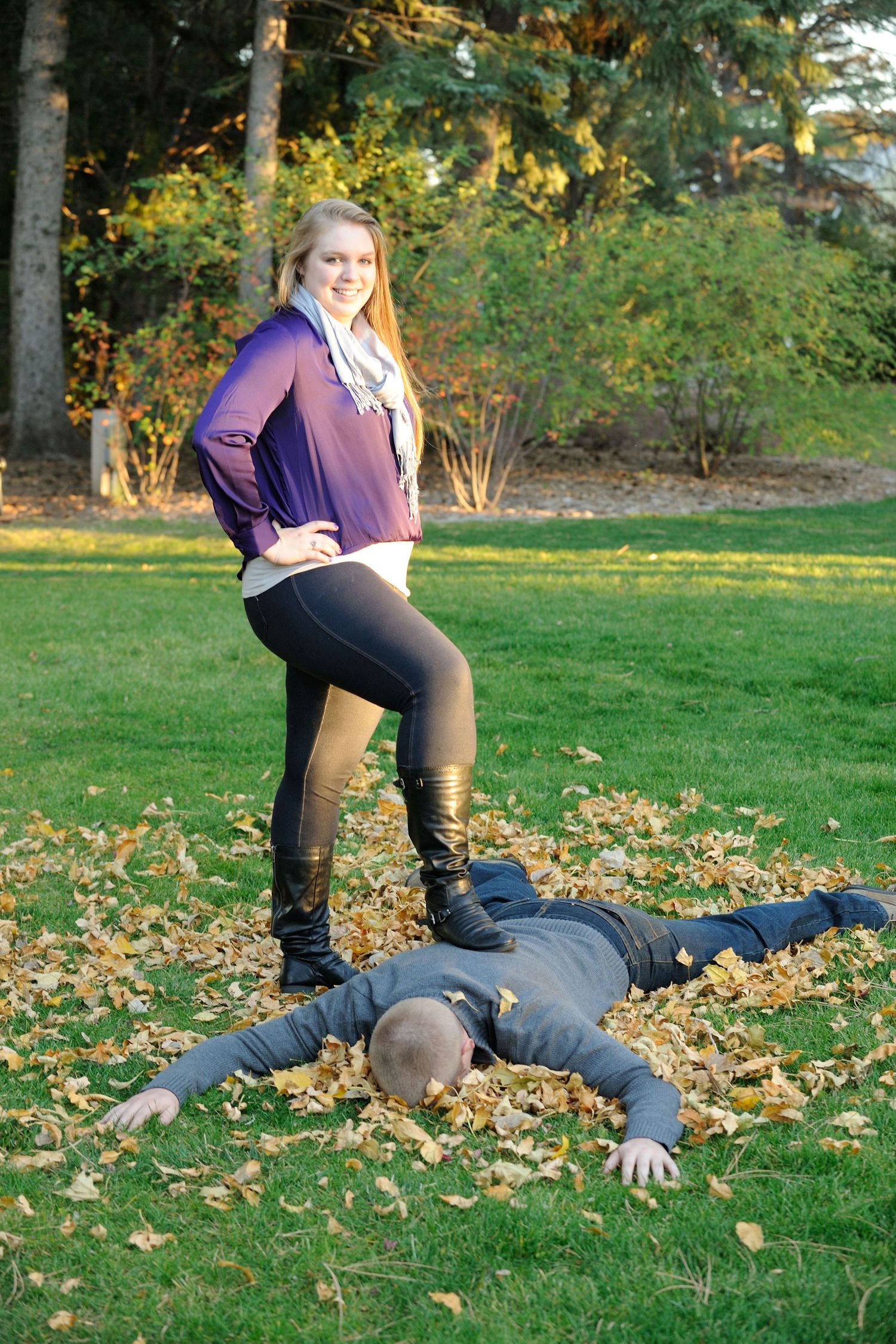 A playful autumn scene showing someone lying in scattered leaves on grass while another stands nearby.