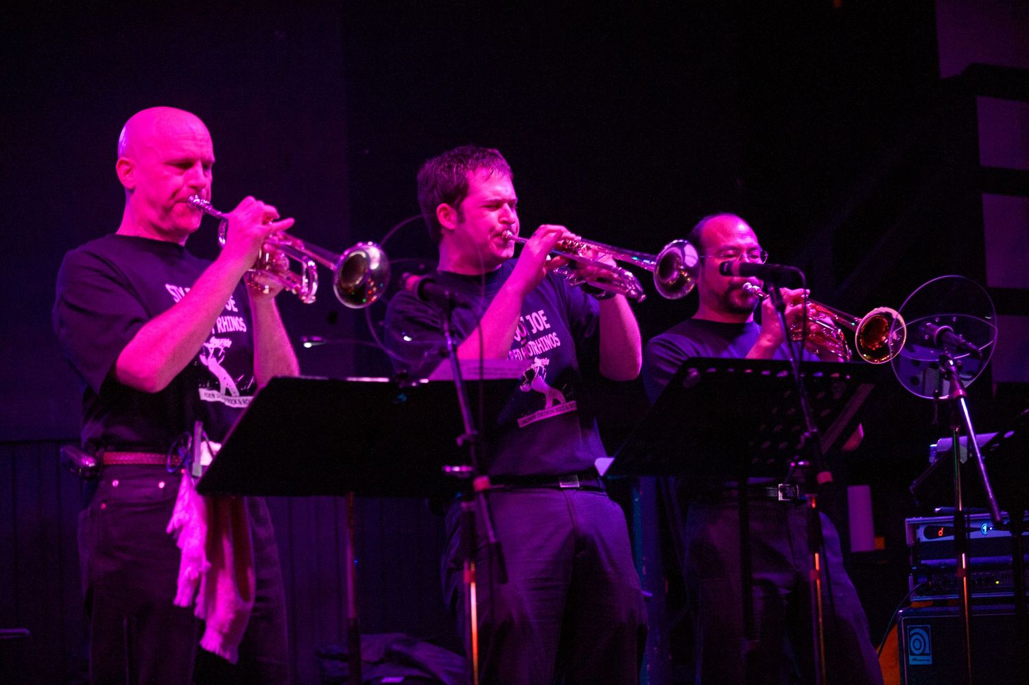 Musicians perform with brass instruments on a darkened stage with pink lighting.