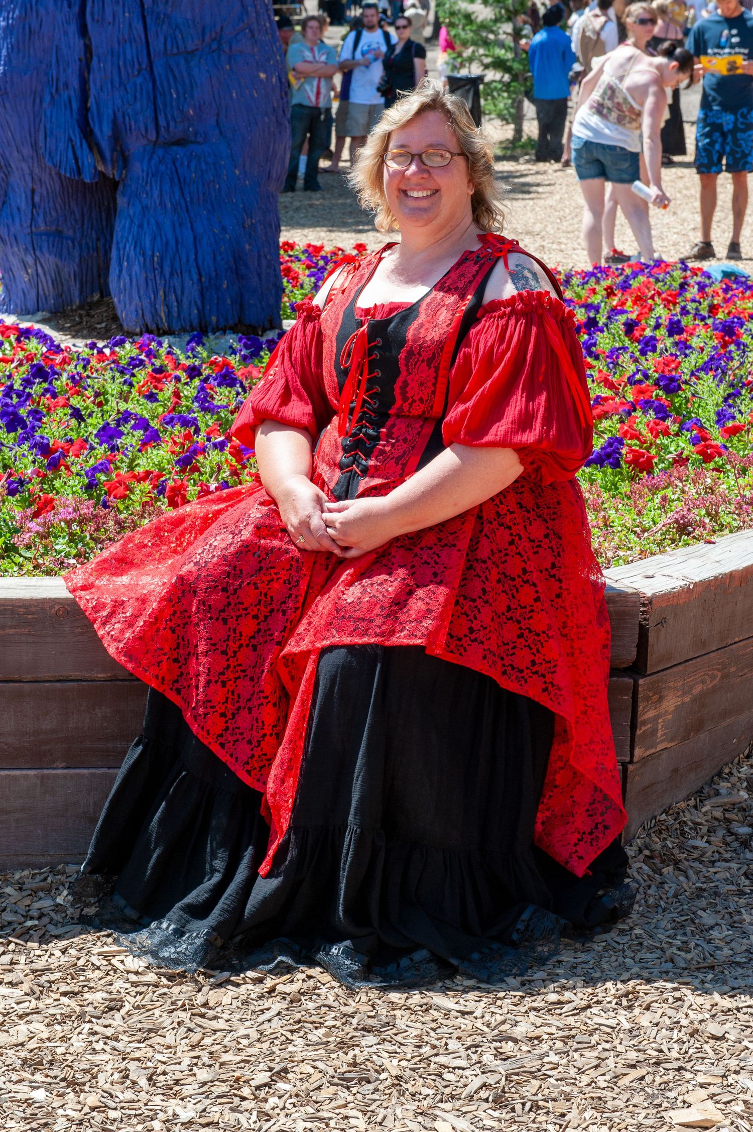 Women in red Renaissance dresses sit on a stone bench near colorful flower beds.