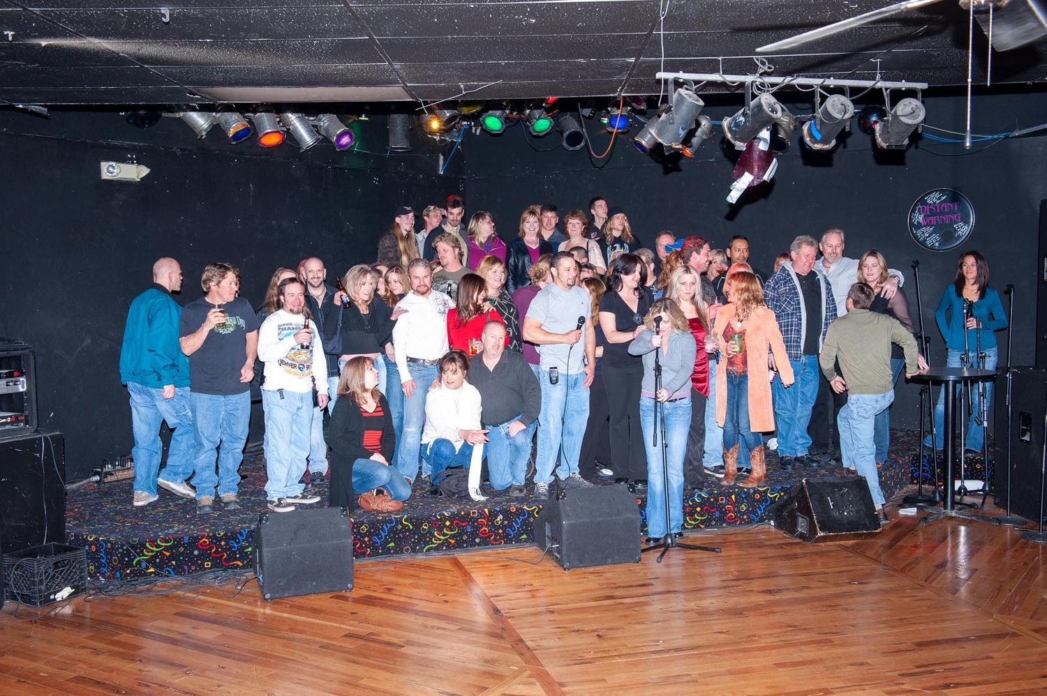 A large group of people standing together on a wooden stage under stage lighting at what appears to be a music venue.