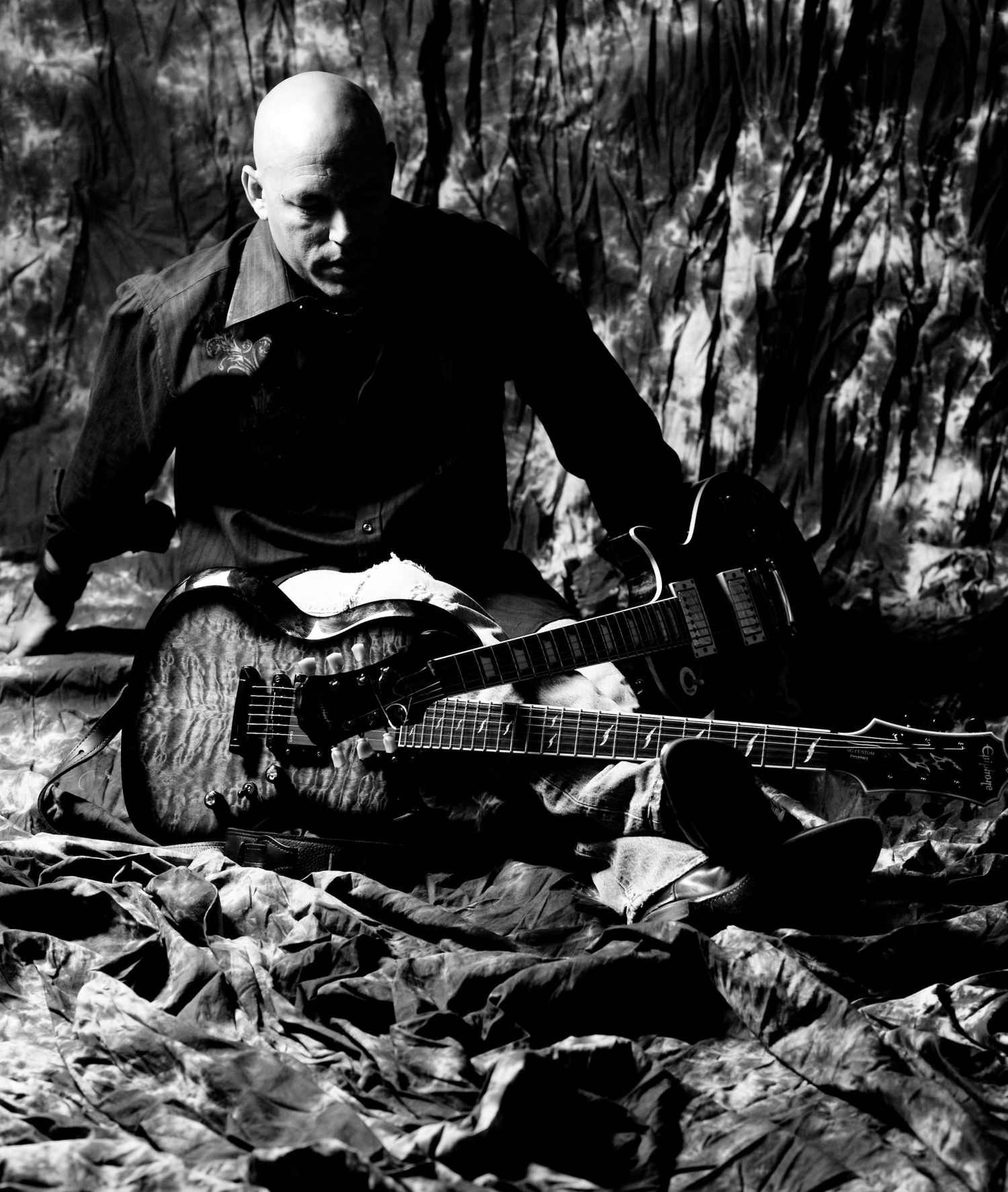 Dramatic black and white photo of a musician with guitars on a textured ground.