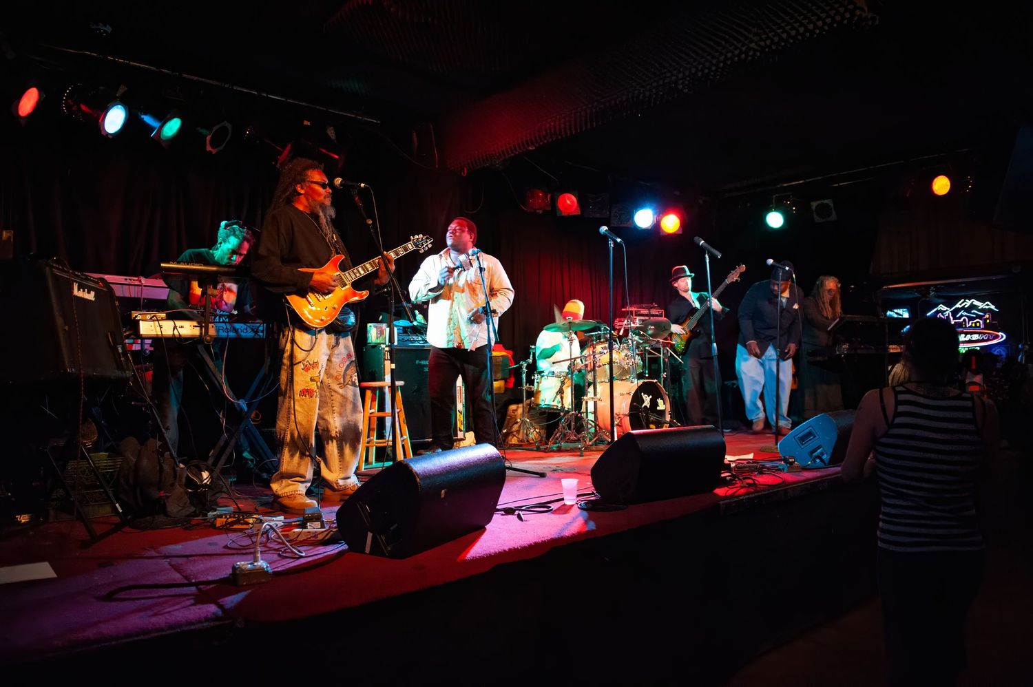 Live band performing on a dimly lit stage with colorful lights, multiple musicians playing guitars and drums in a music venue.