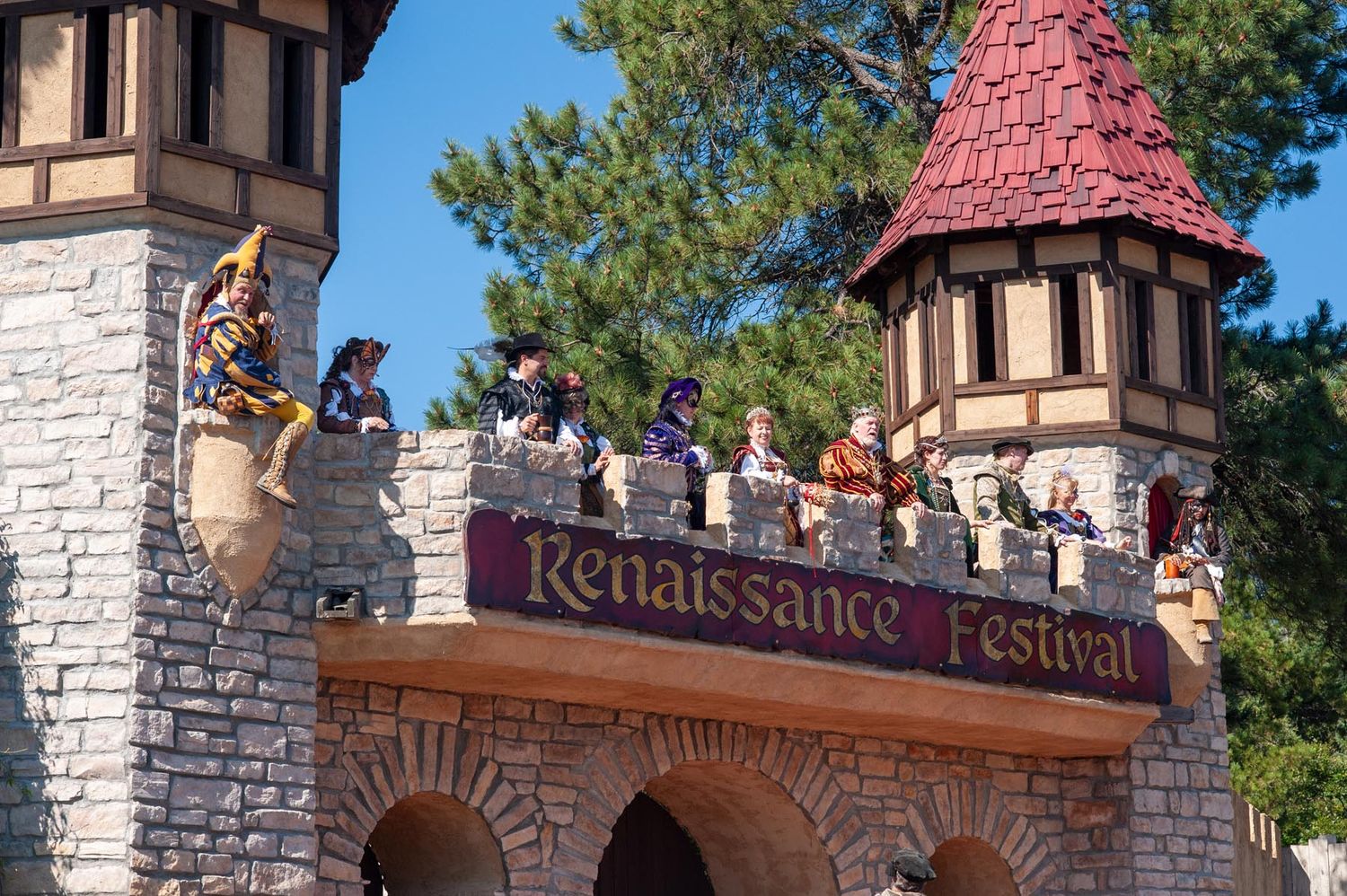 Stone entrance gate with red turrets and decorative signage welcomes visitors to a Renaissance festival.
