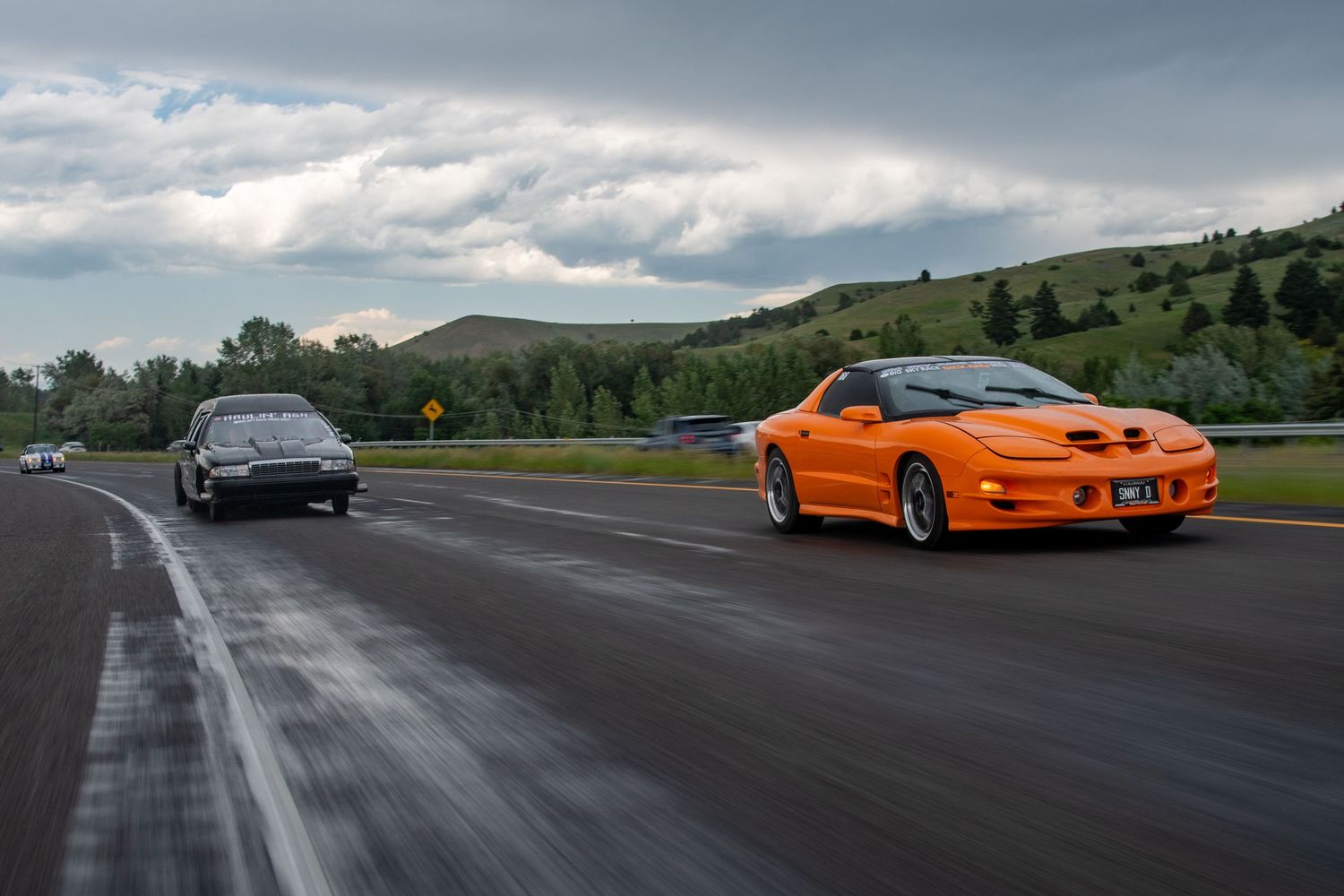 orange Pontiac firebird on the right and drag racing converted hearse driving on the left with beautiful mountains in the background
