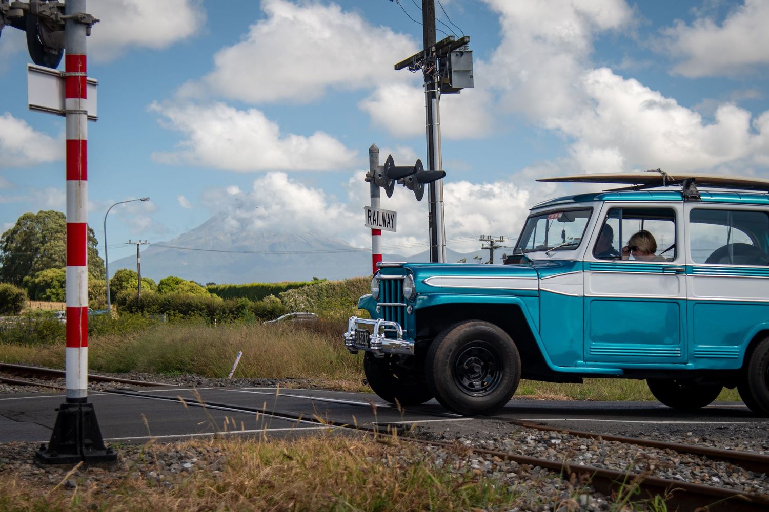 blue and white jeep crossing railroad tracks with mount Taranaki New Zealand in the background