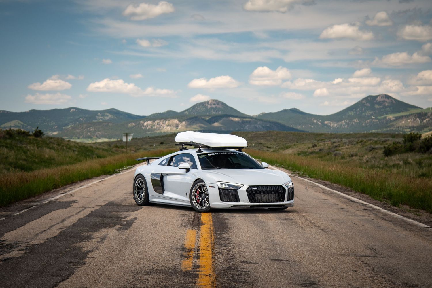 White Audi R8 with roof cargo box, custom wheels, and dark window tint in beautiful landscape with mountains off in the distance.