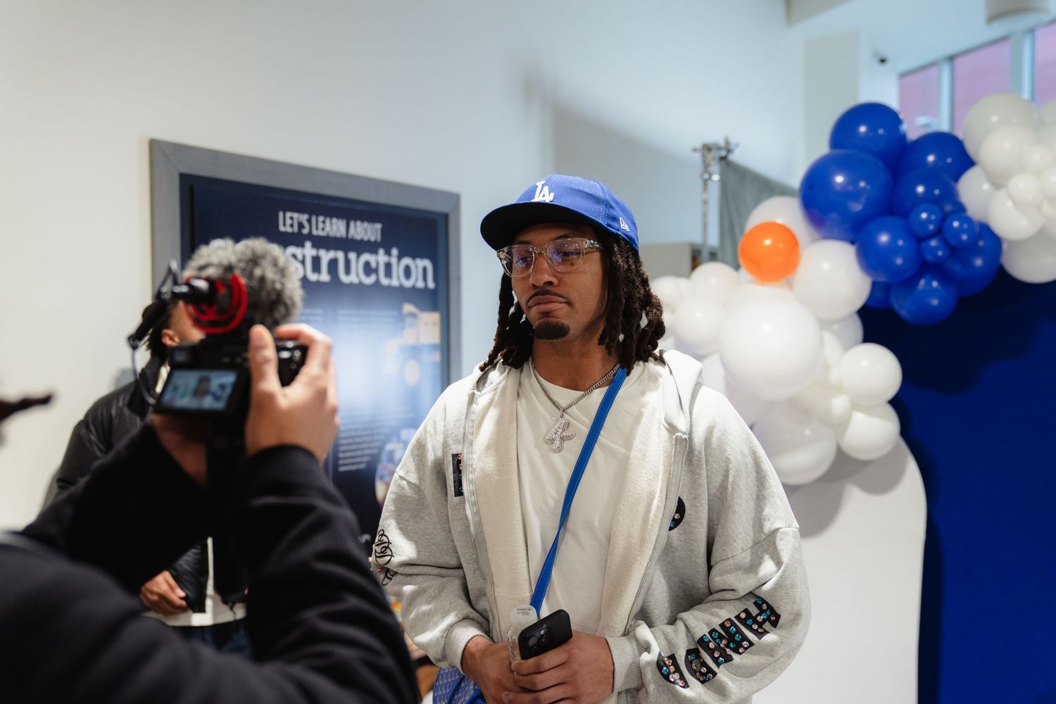 Young man in white jacket and blue cap being filmed at an indoor community event with balloons.
