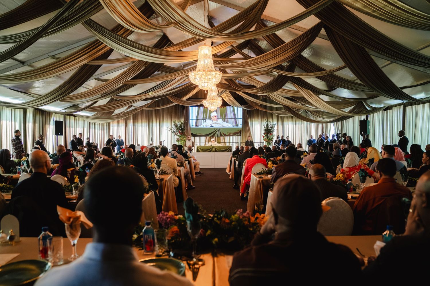 Large formal banquet hall with draped ceiling, chandelier, and guests seated at decorated tables.