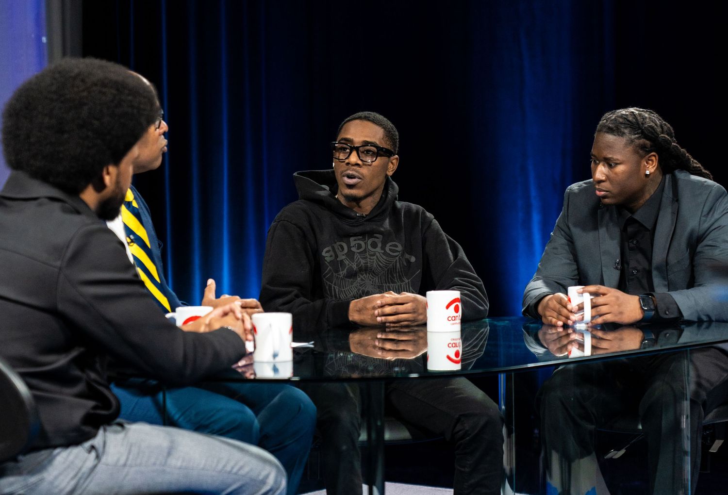 Three men engaged in discussion around a glass table in a TV studio setting.