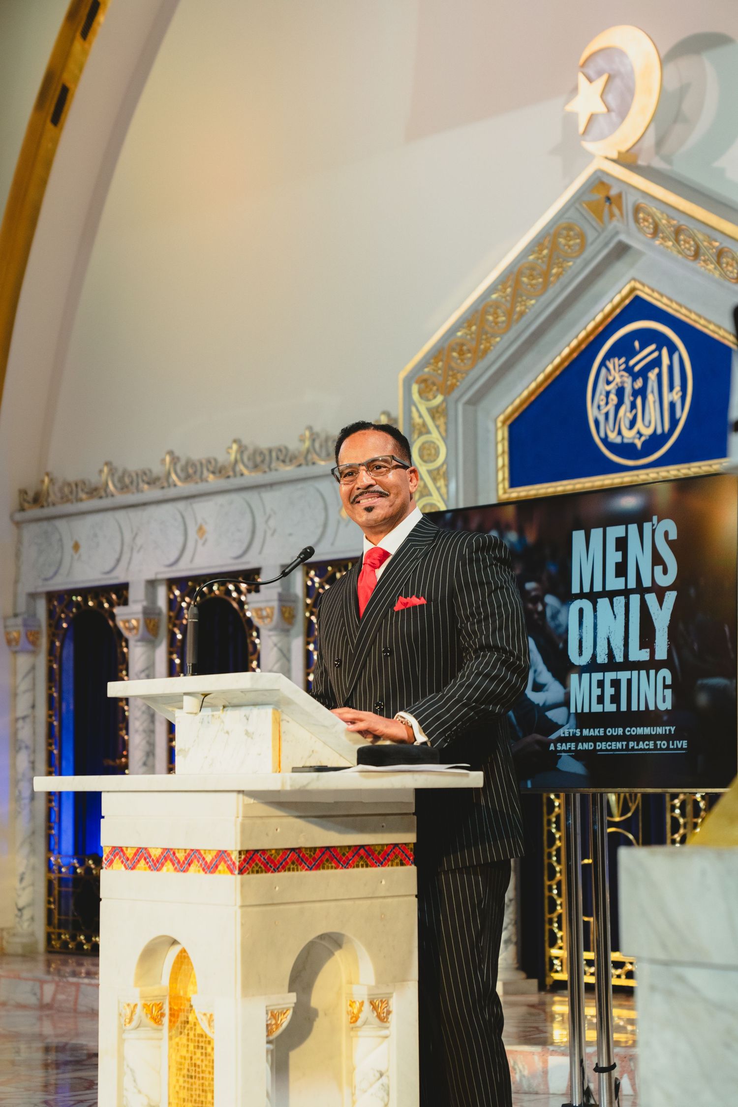Man in pinstripe suit speaks at podium during Mens Only Meeting at mosque.