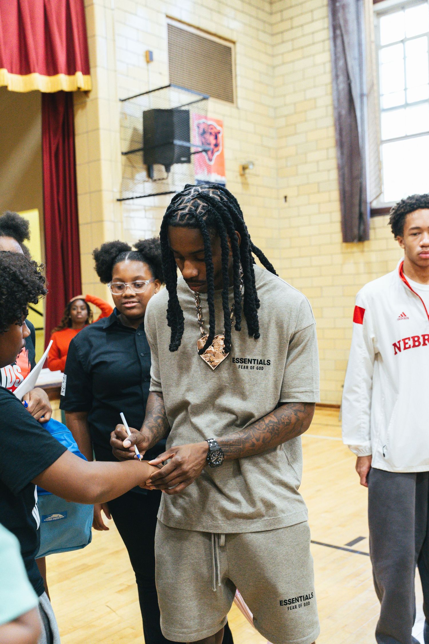 A young man with dreadlocks wearing a beige Essentials tracksuit signs autographs at a school gymnasium event.