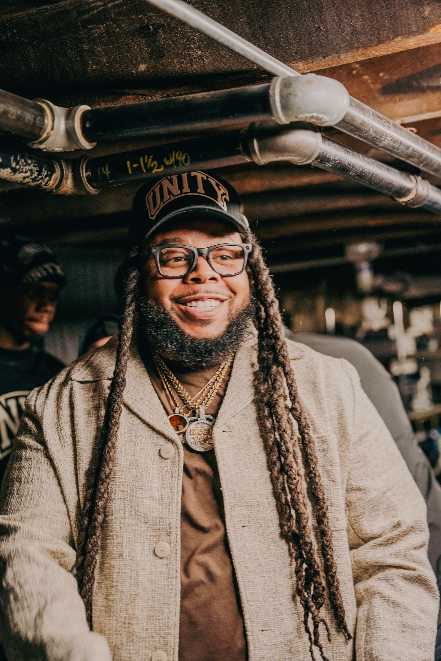 Smiling man with long dreadlocks wearing Unity cap and glasses indoors.