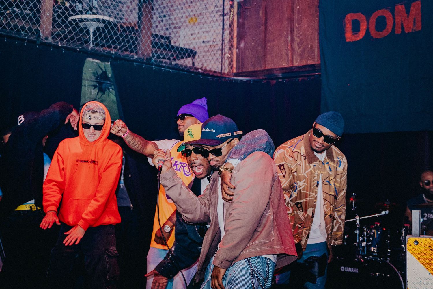 Group of young men in colorful streetwear posing together at an outdoor urban music event at night.