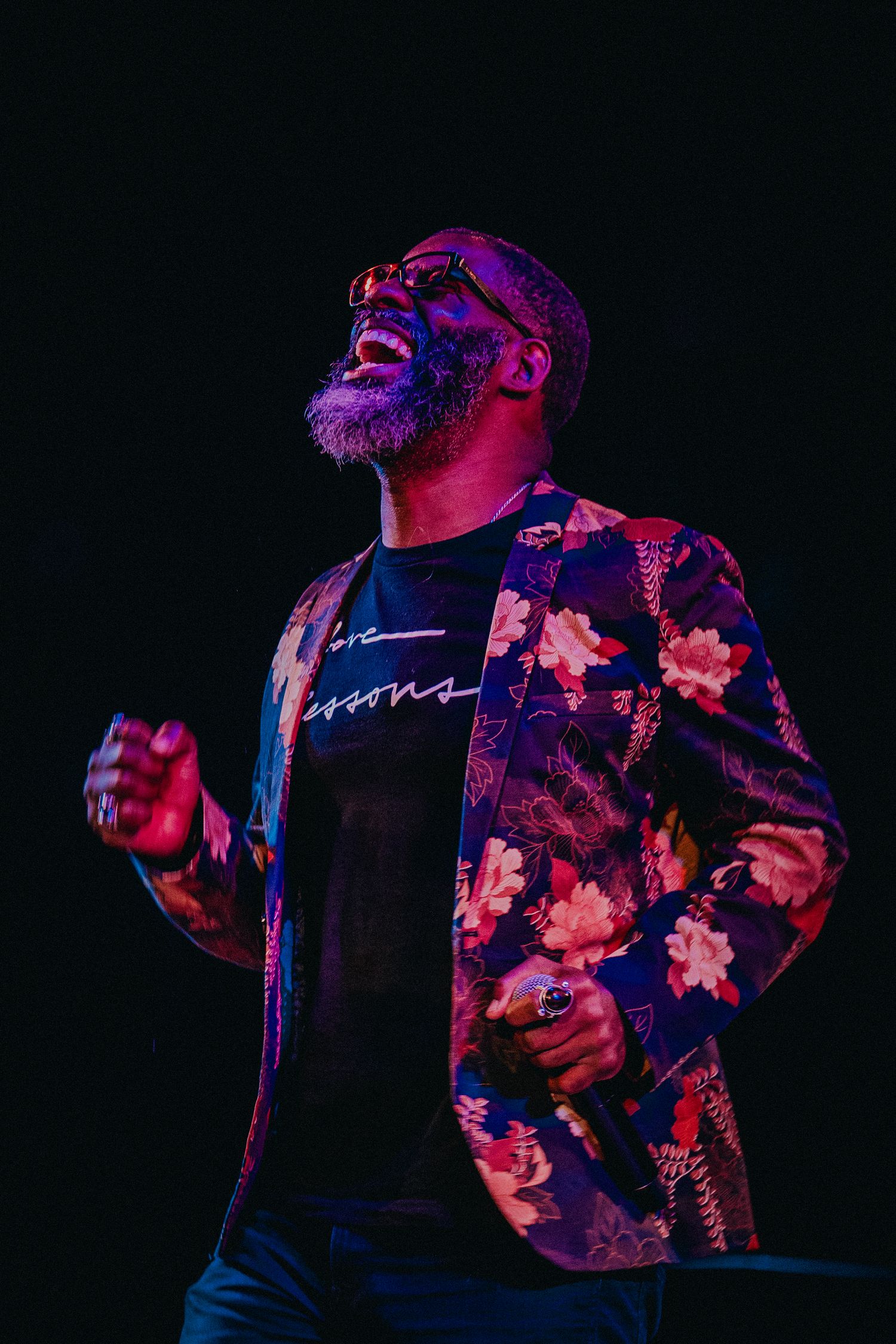 A man in a floral blazer and sunglasses performs passionately on stage under purple and blue stage lighting.