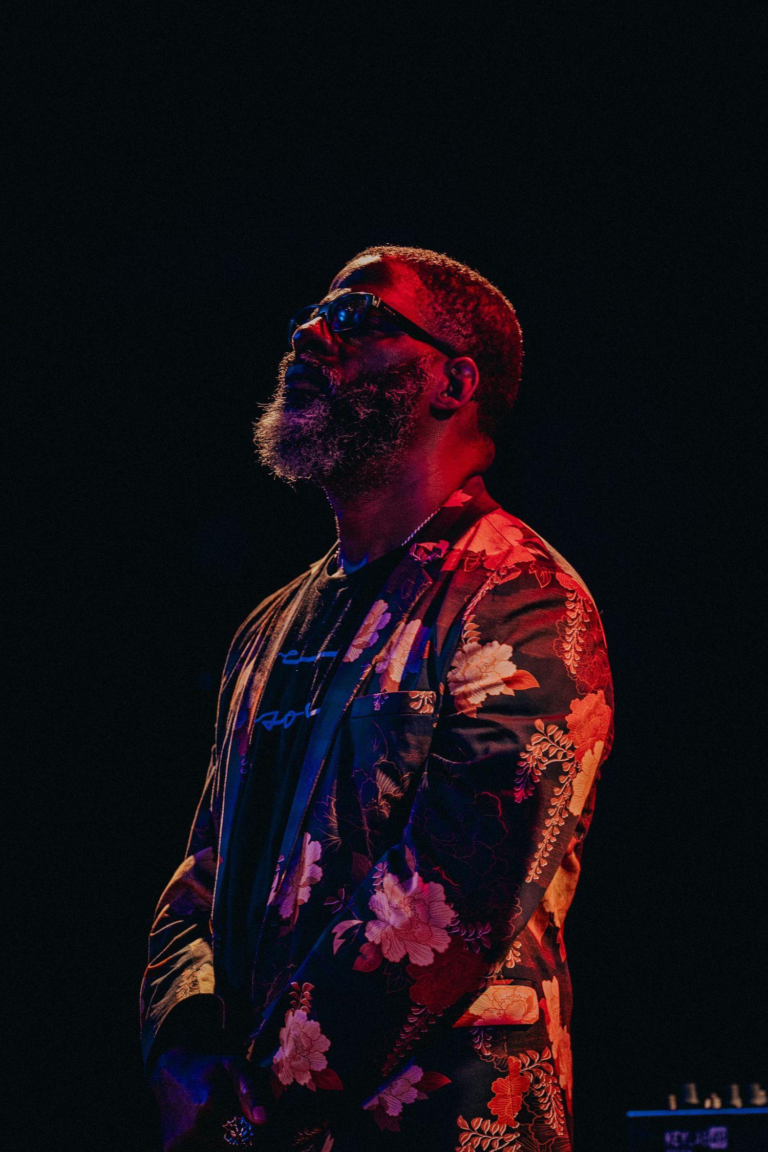 Man in floral jacket and sunglasses performing on stage under dramatic red and blue lighting.