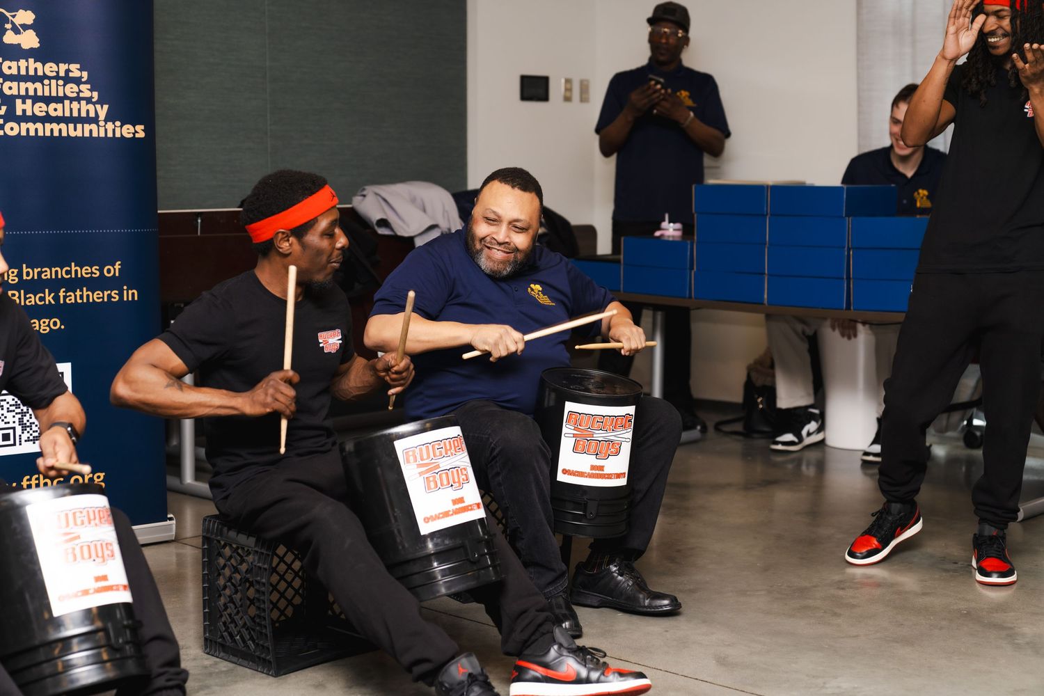 Two men smiling and playing bucket drums at a community organization event indoors.