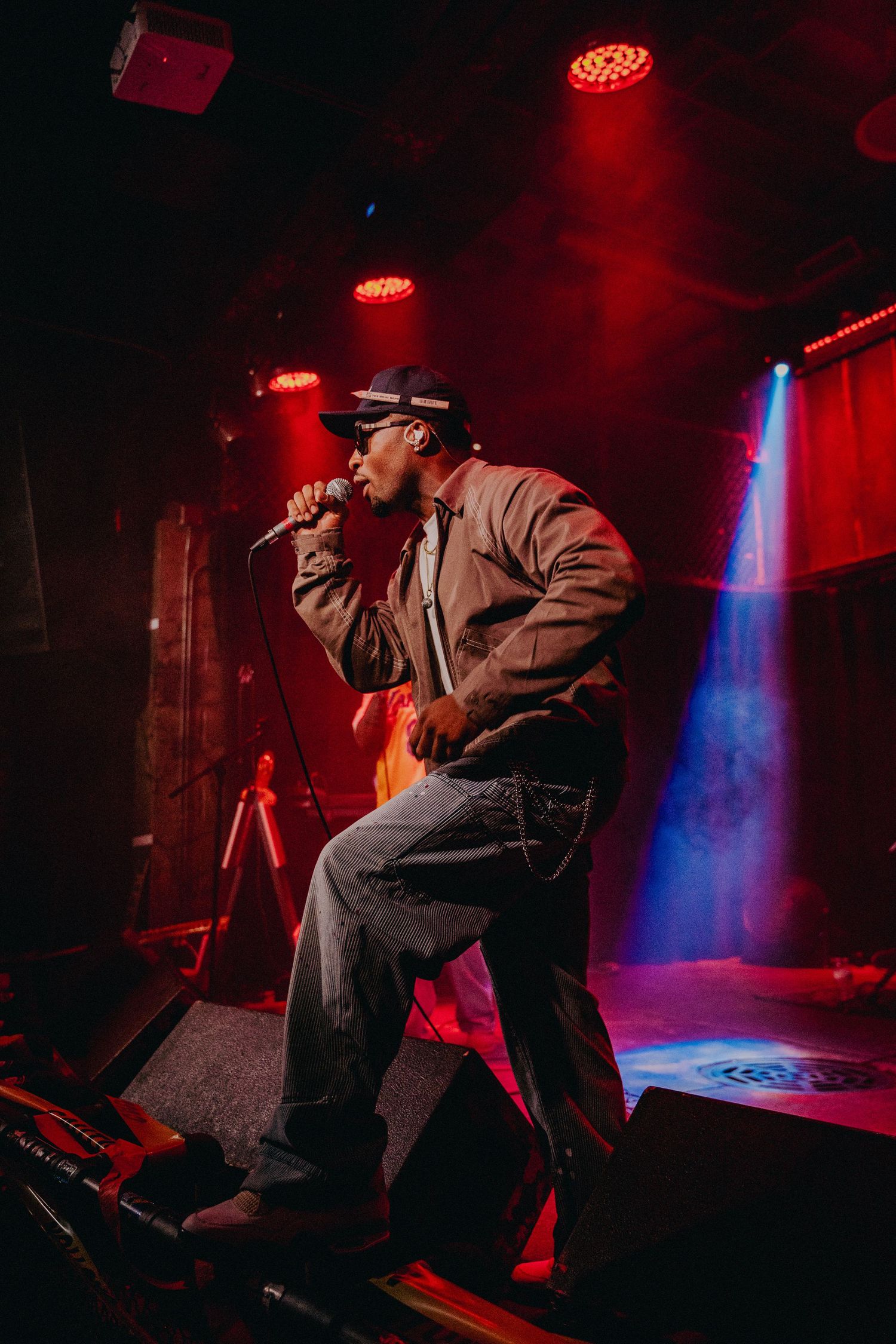 Hip-hop artist performing on stage under red and blue stage lights at an indoor music venue.