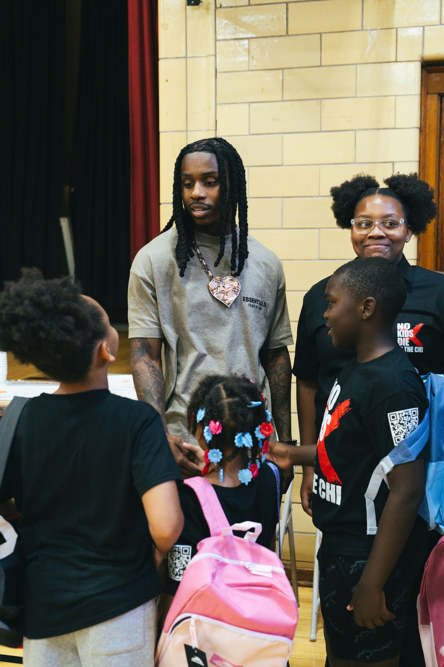 A tall man with dreadlocks and a heart pendant necklace interacts with children carrying backpacks at a community event.