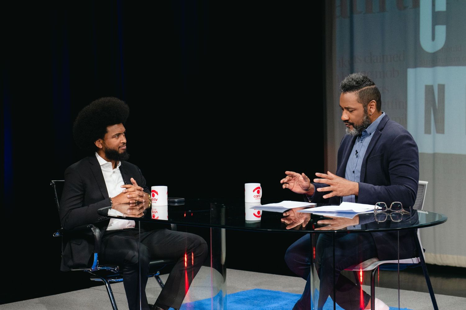 Two men in conversation at a professional interview setup on a dark stage.