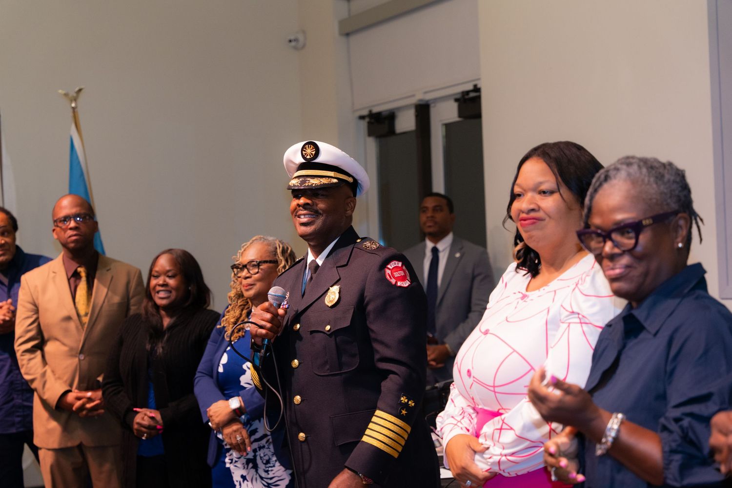 A Black fire chief in uniform stands smiling with community members at an official recognition ceremony.