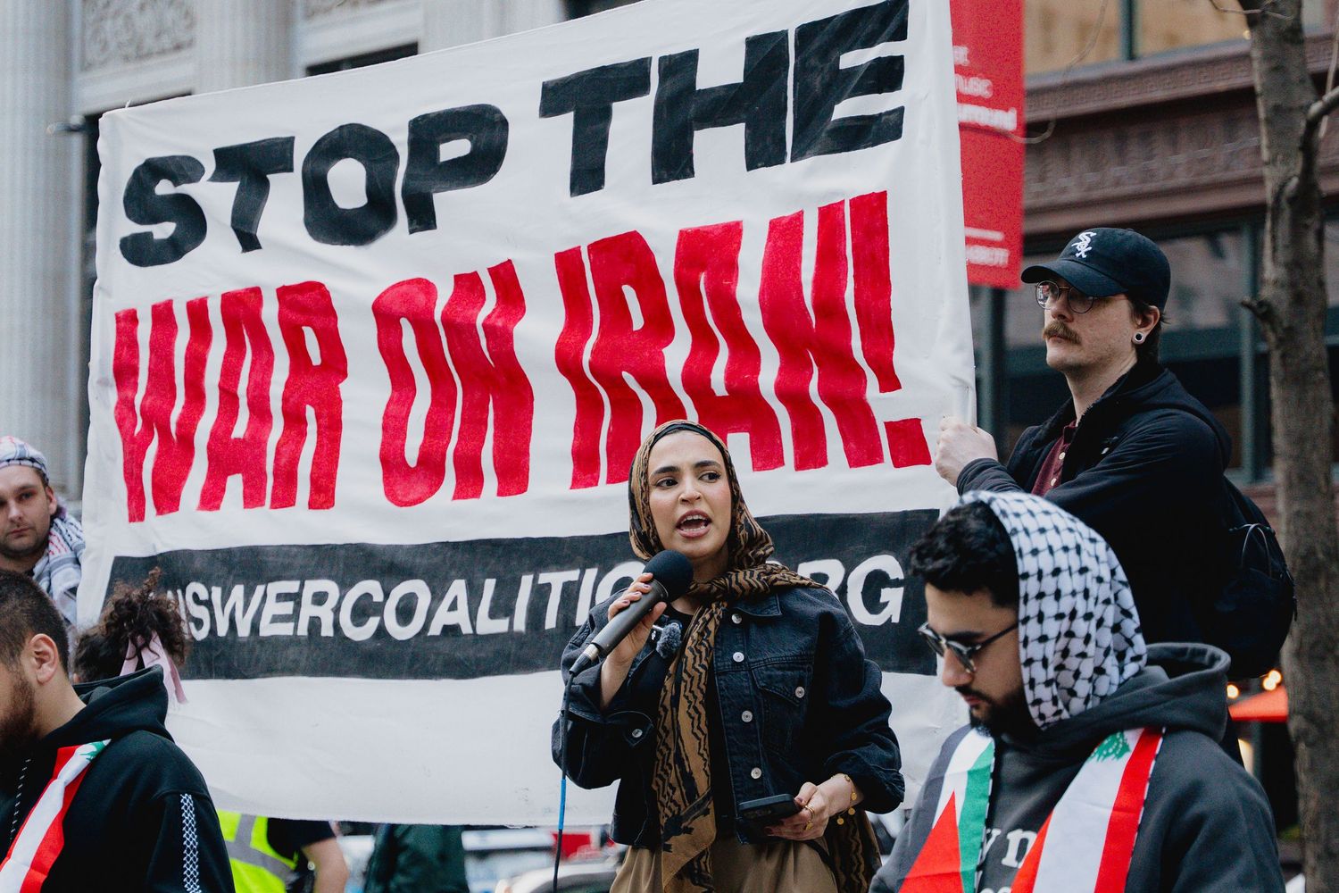 Protesters hold Stop the War on Iran banner at outdoor demonstration.