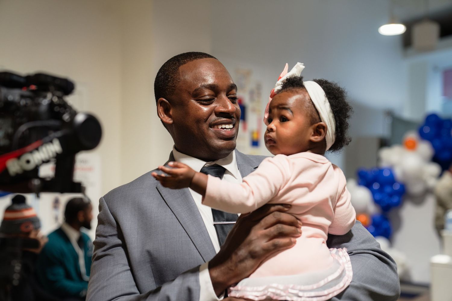 A man in a gray suit smiles while holding a baby girl wearing a pink outfit and white bow headband.