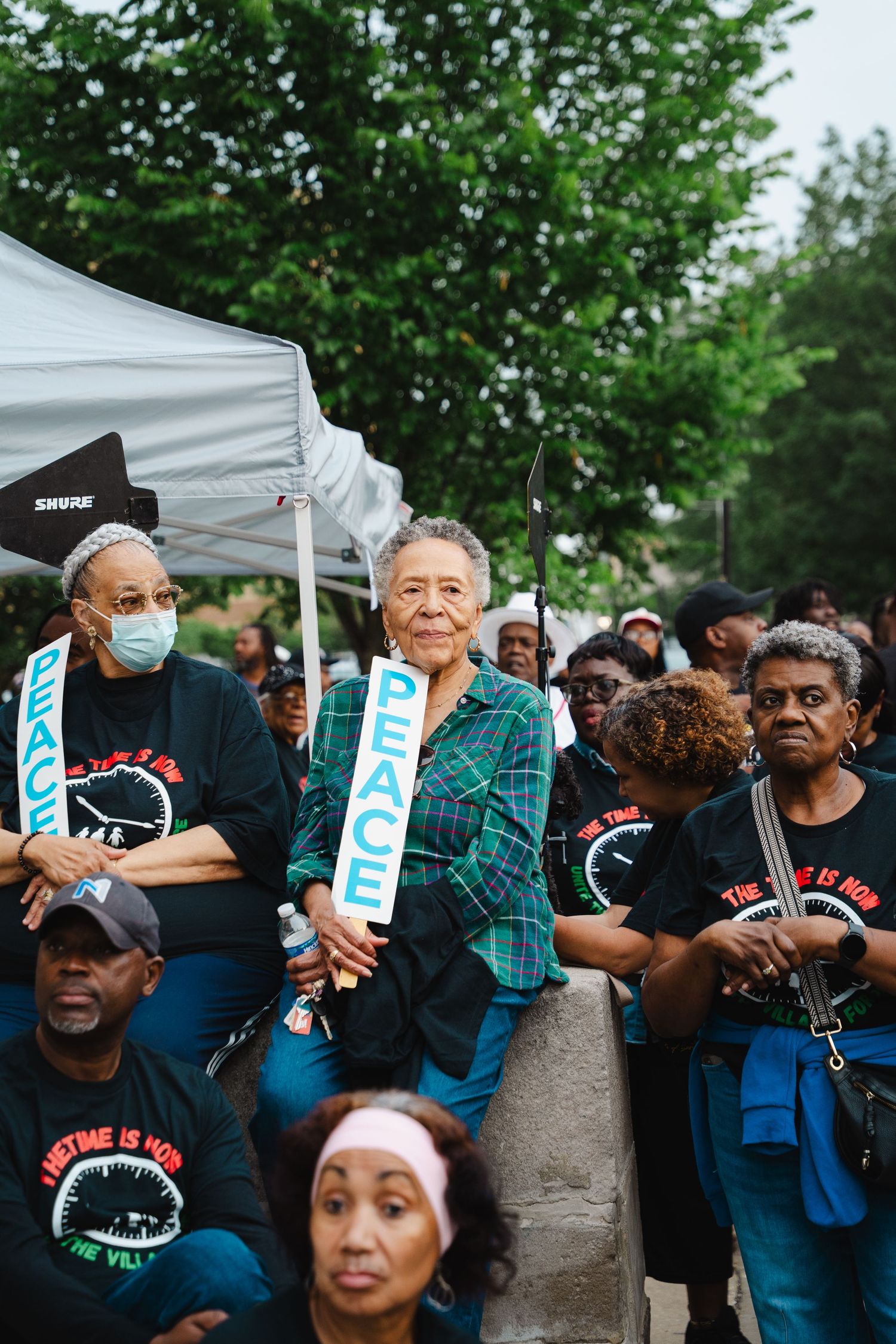 Crowd of community members in matching black shirts holding peace signs at an outdoor rally.