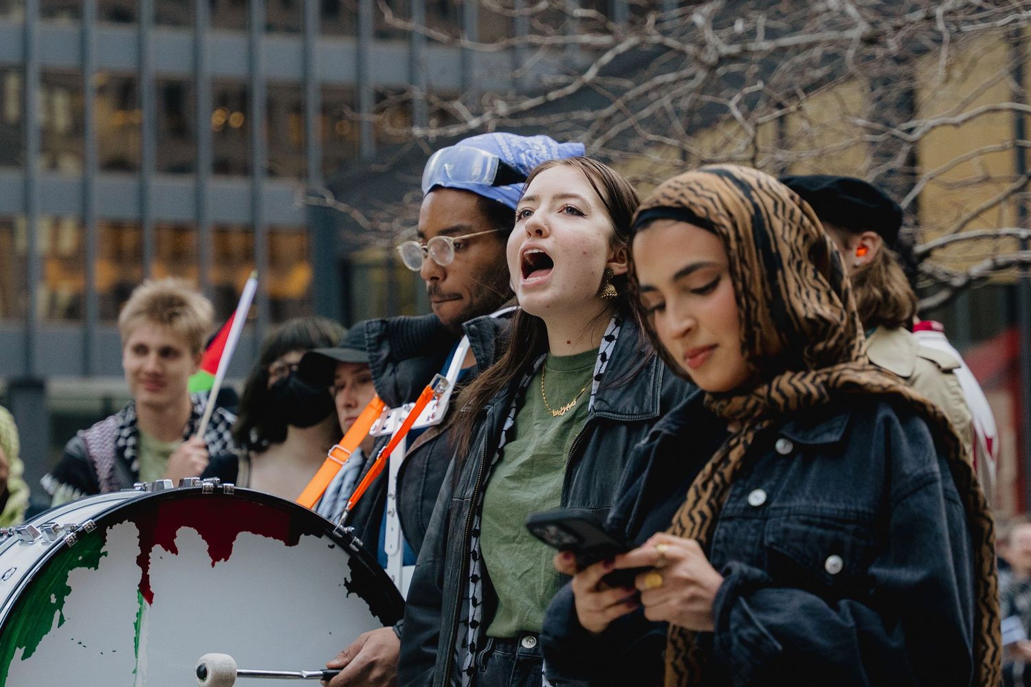 Young woman chants at outdoor protest rally with drum and crowd behind her.