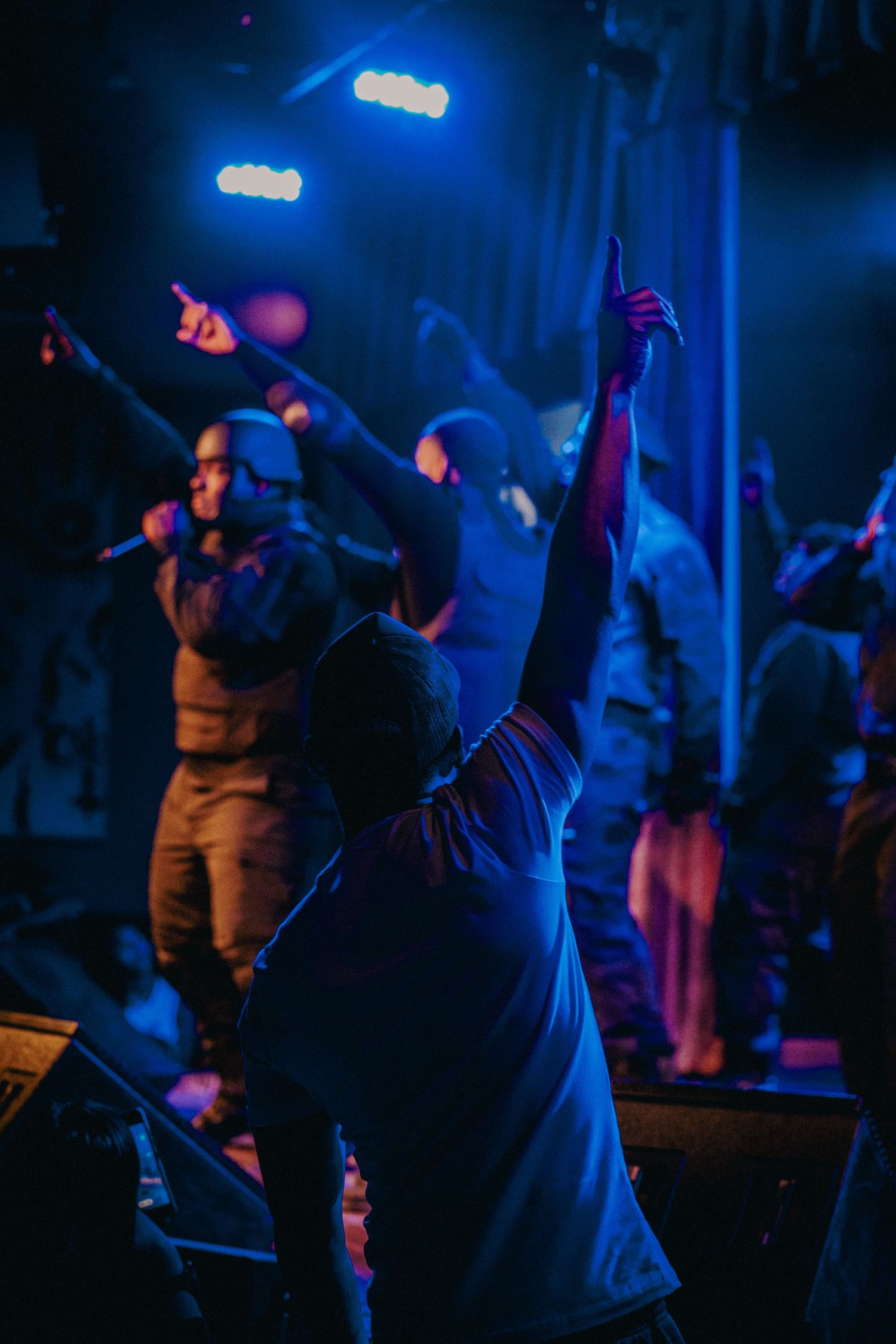 Performers on stage with blue stage lighting as an audience member raises their hand at a live concert.