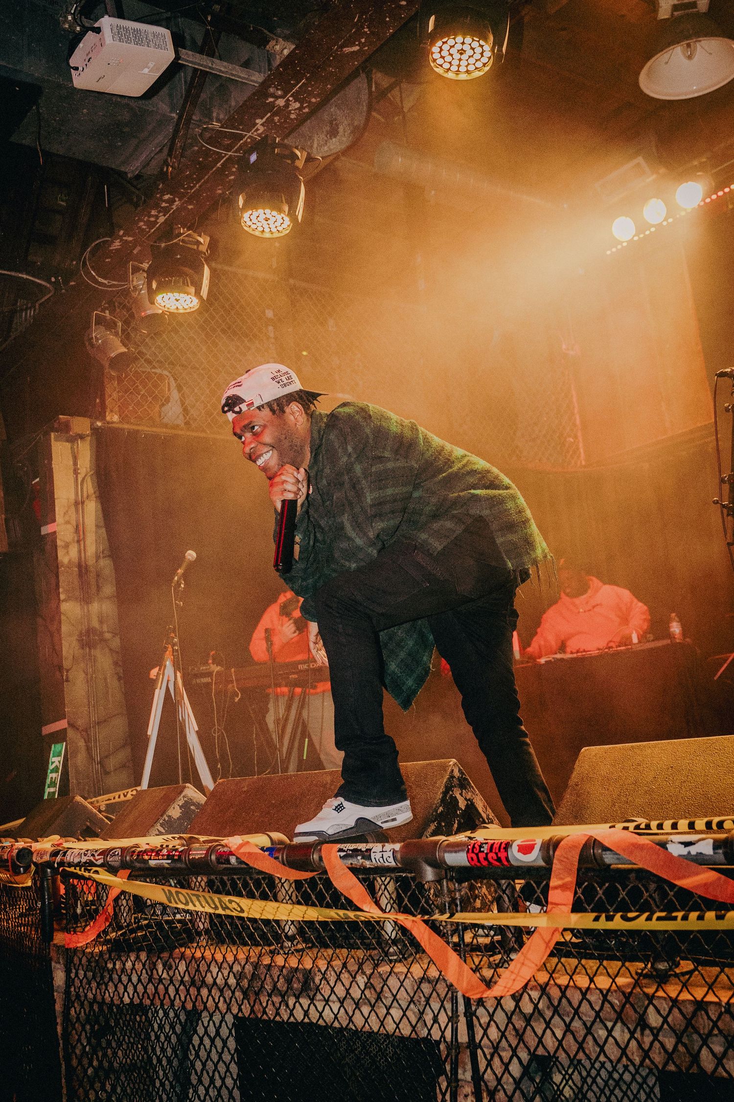 Male rapper leaning into microphone on stage under warm golden stage lights at a live show.
