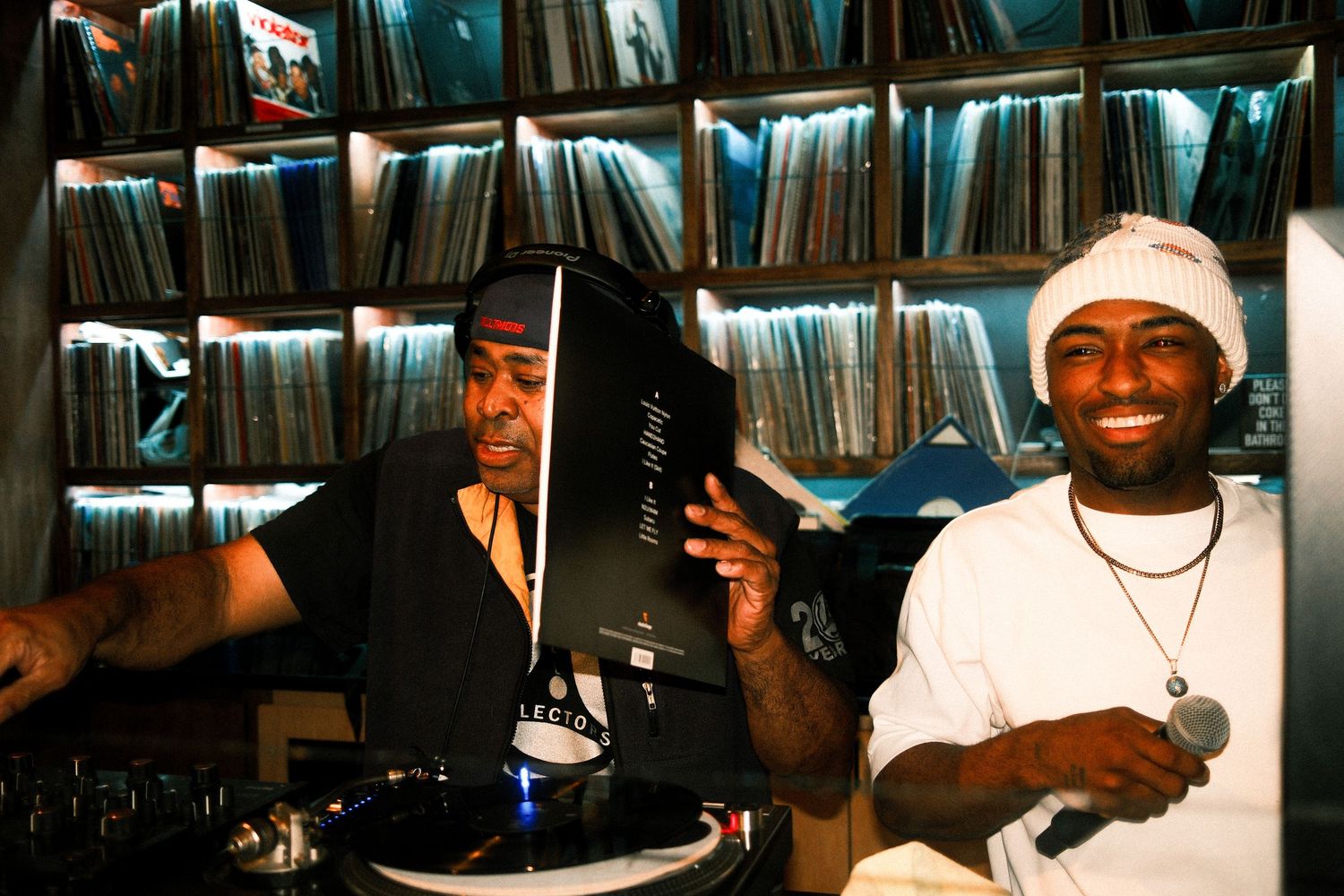 Two DJs smile while working turntables surrounded by vinyl record collection.
