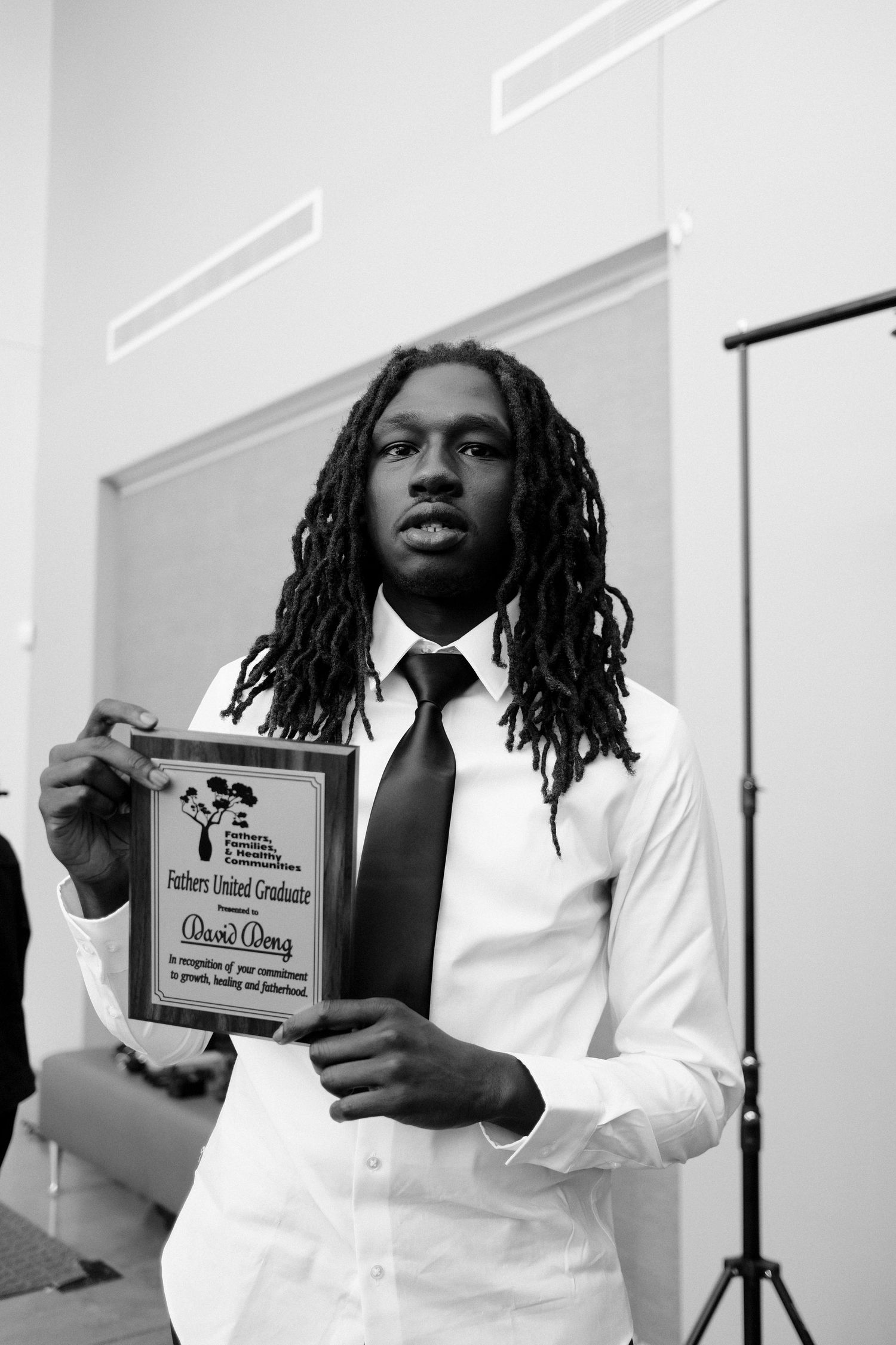 Young man with dreadlocks holding a Fathers United Graduate award plaque in black and white photo.