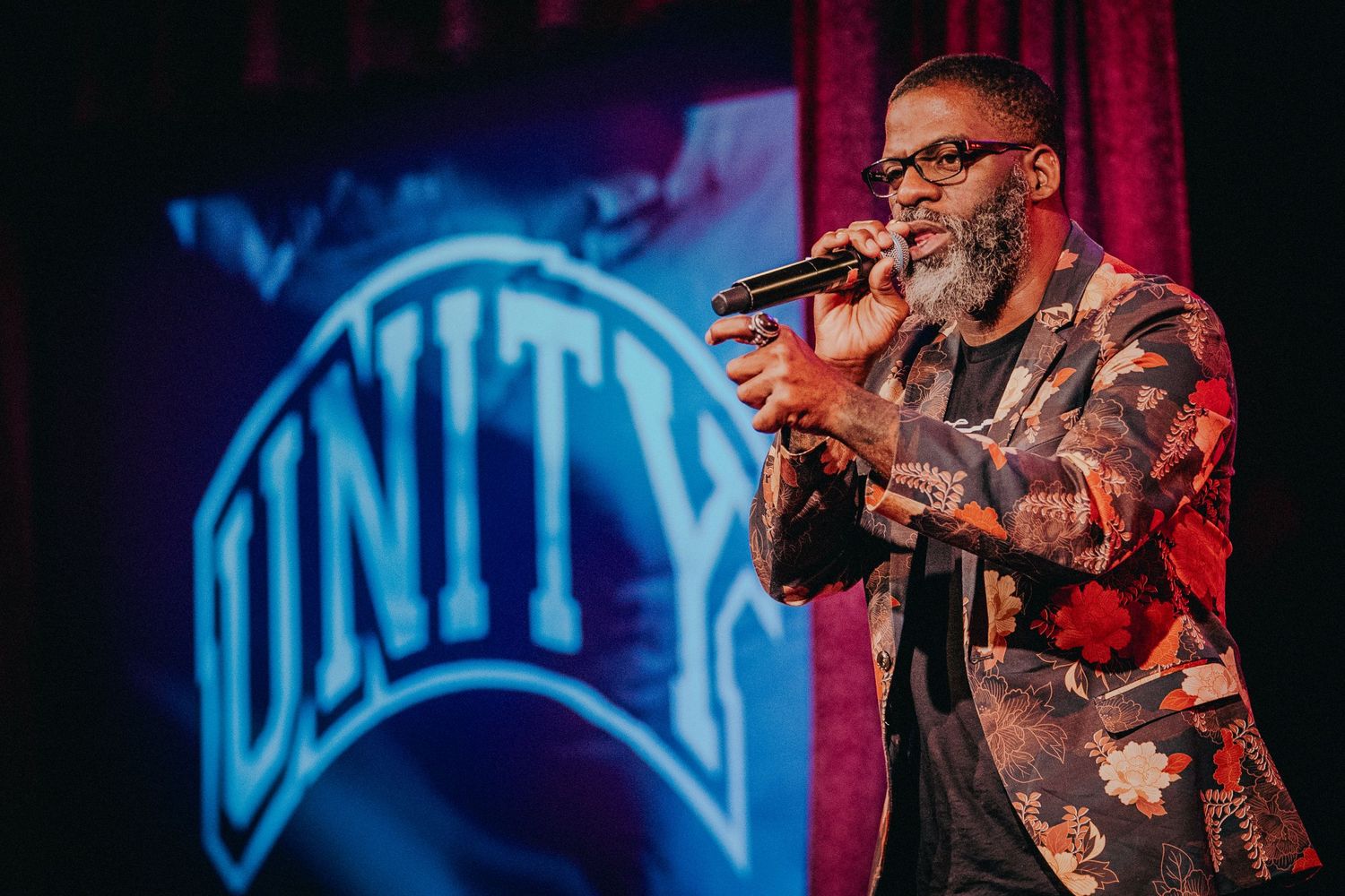 A performer in a floral jacket holds a microphone on stage with a Unity logo projected in the background.