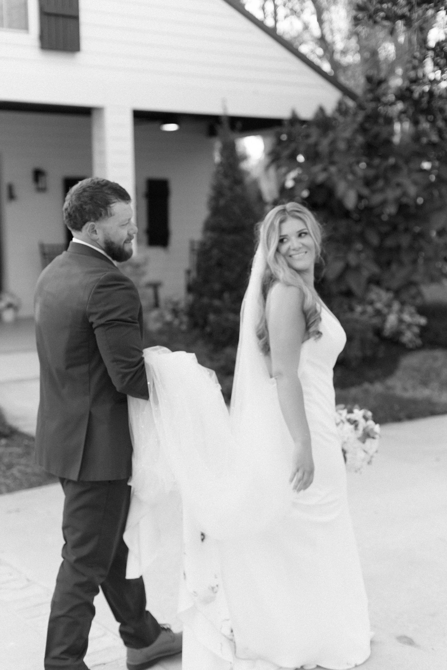 Black and white wedding photo of a couple with a flowing veil outside a white house.