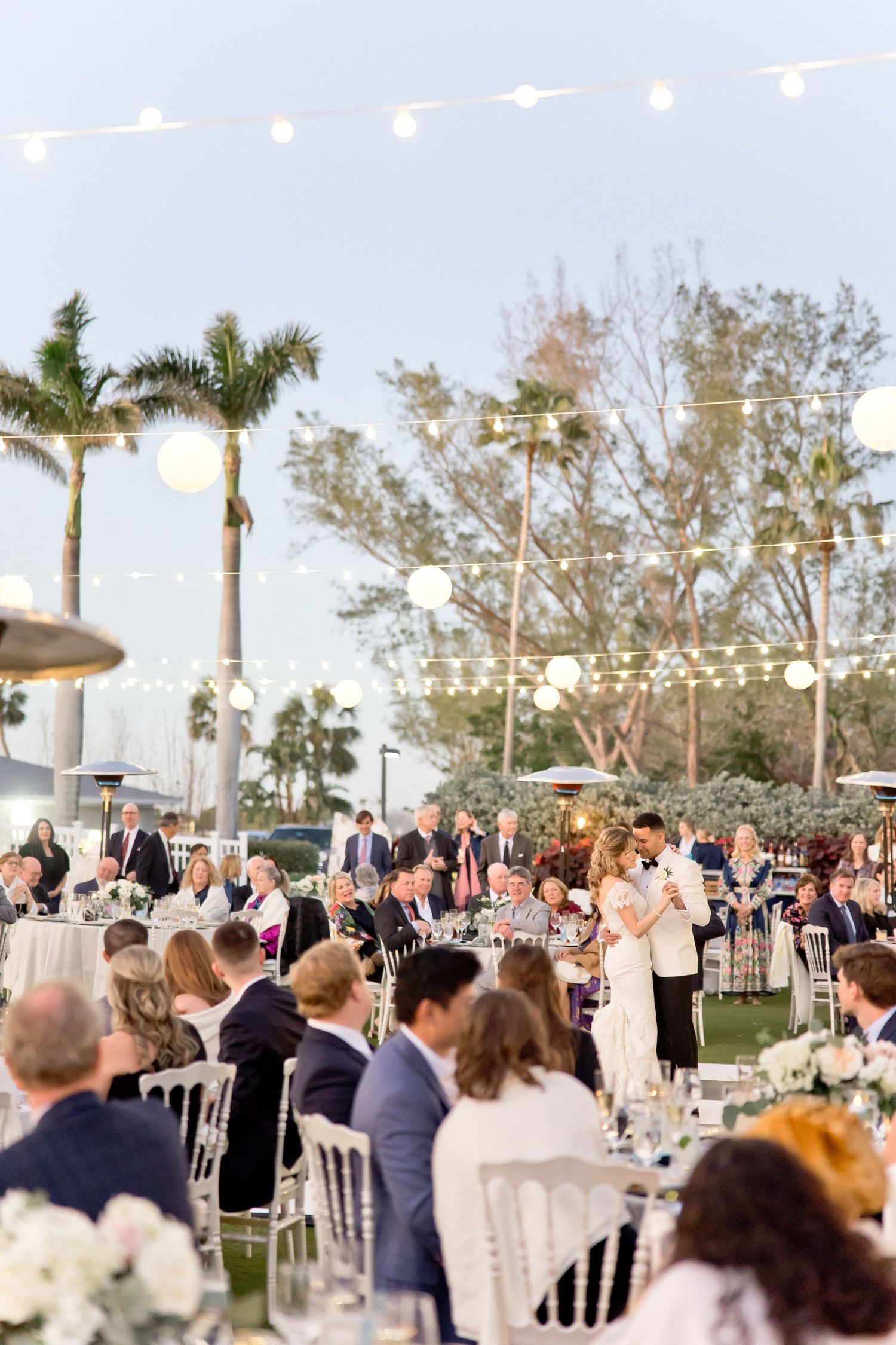 An outdoor wedding reception with string lights and palm trees features guests dining at elegant tables.