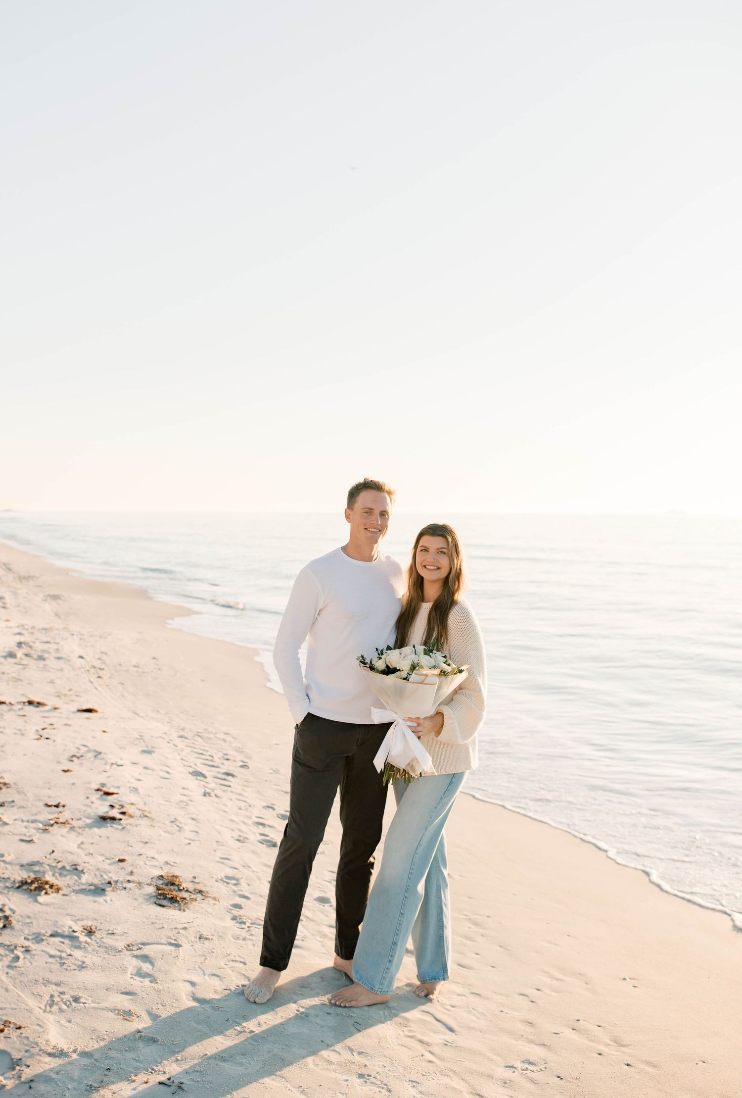 A couple in coordinated white outfits poses together on a sandy beach at sunset.