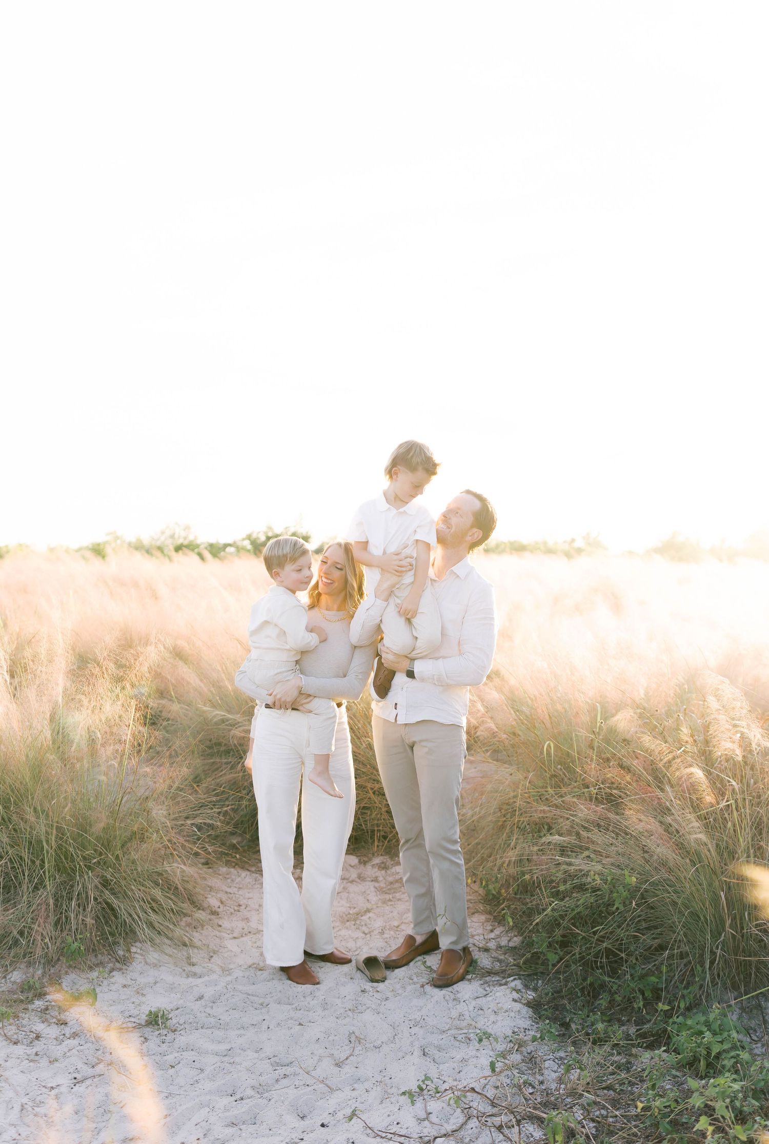 Family portrait in white outfits standing together on a path through tall grass at sunset.