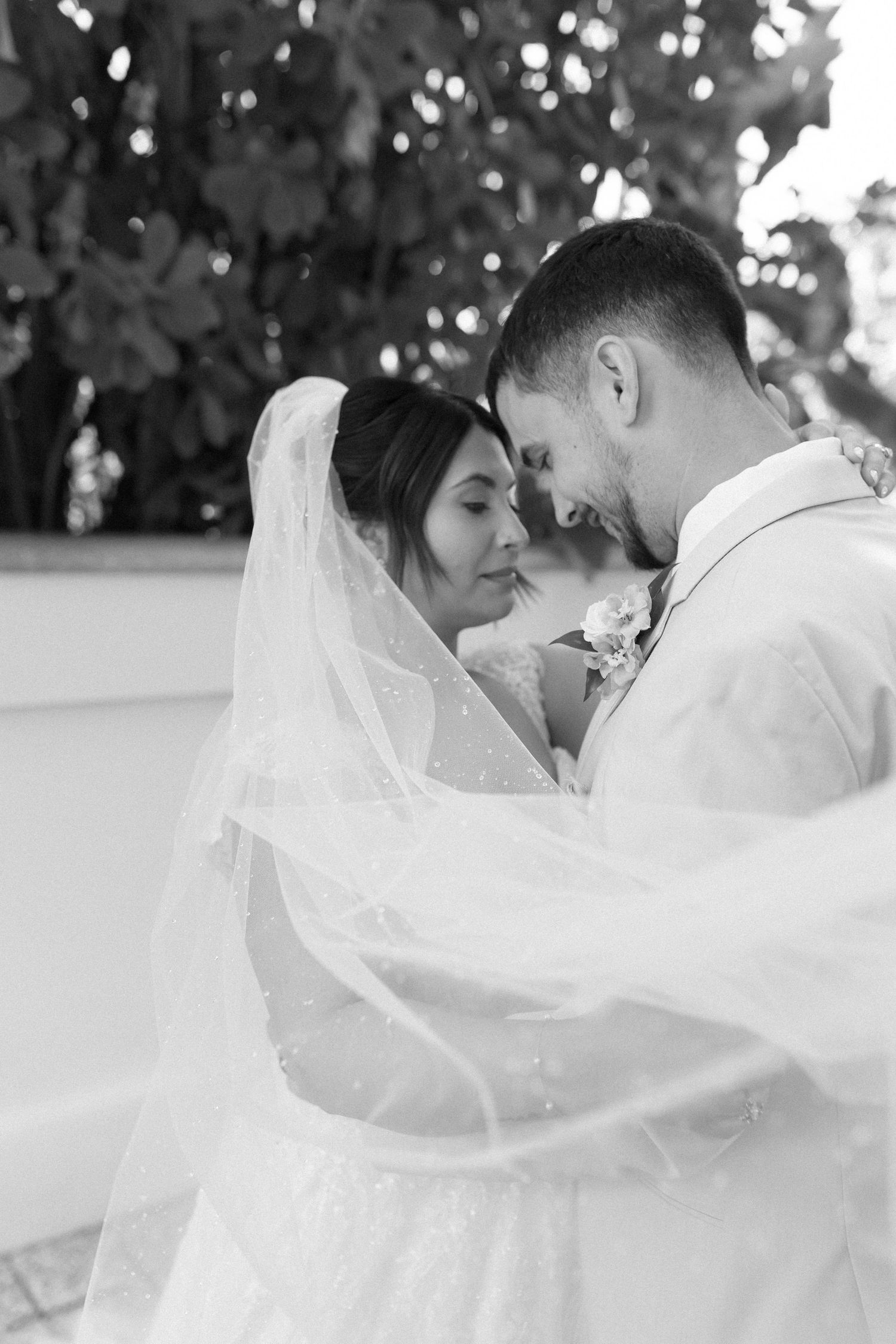 Black and white wedding photo of bride and groom sharing an intimate moment, veil flowing, surrounded by lush greenery.