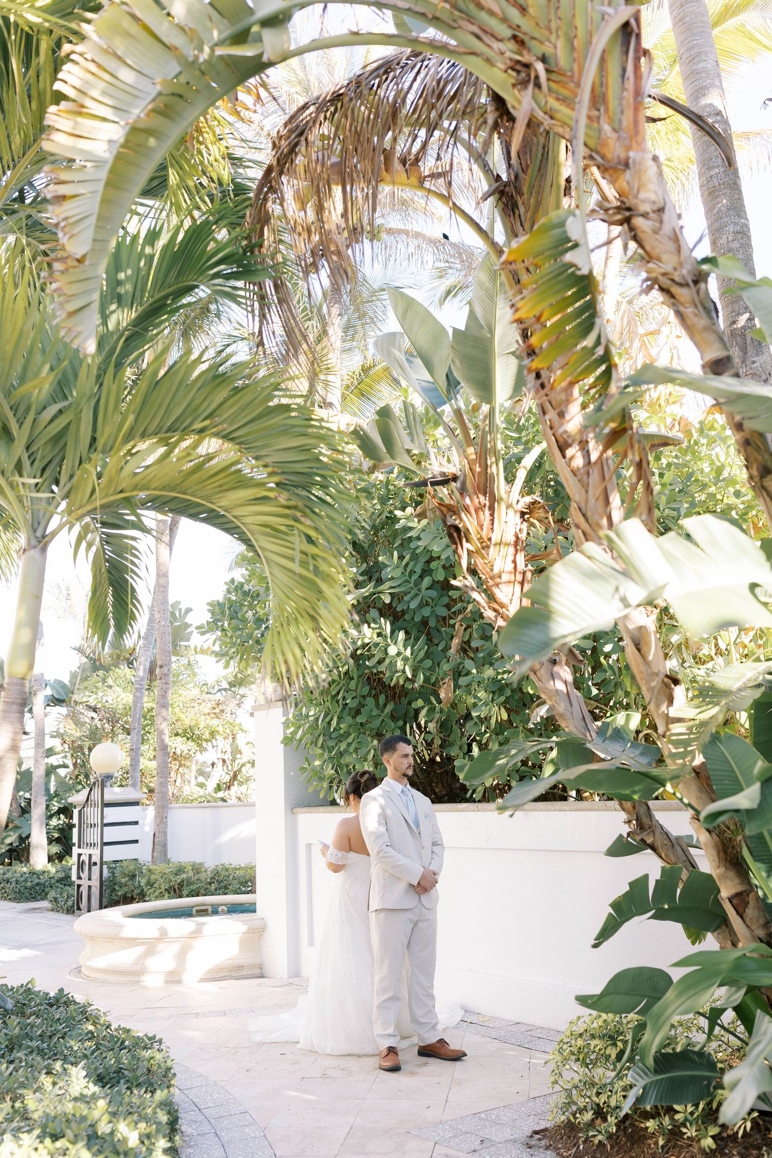 Groom in white linen suit stands among lush tropical palm trees and greenery at an outdoor wedding venue.
