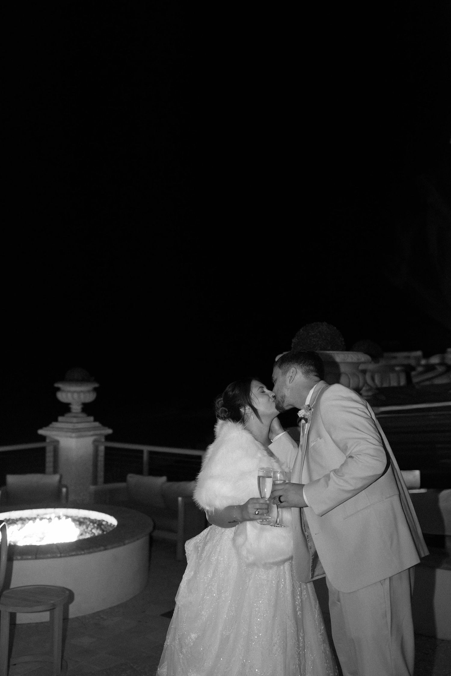Black and white photo of a bride and groom sharing a kiss outdoors at night near a fire pit.