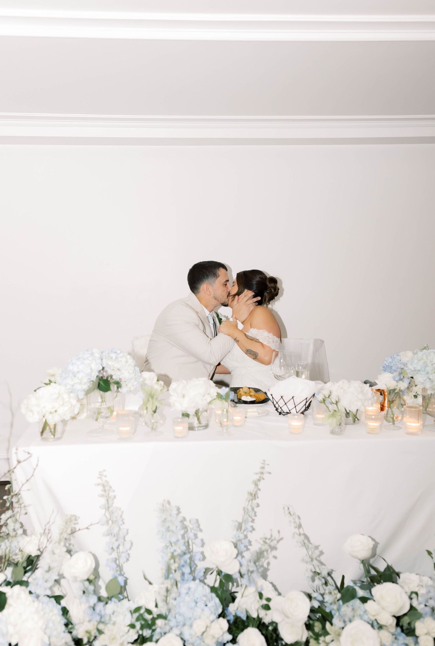 Newlyweds kiss at sweetheart table adorned with blue hydrangeas, white roses, and glowing candles.