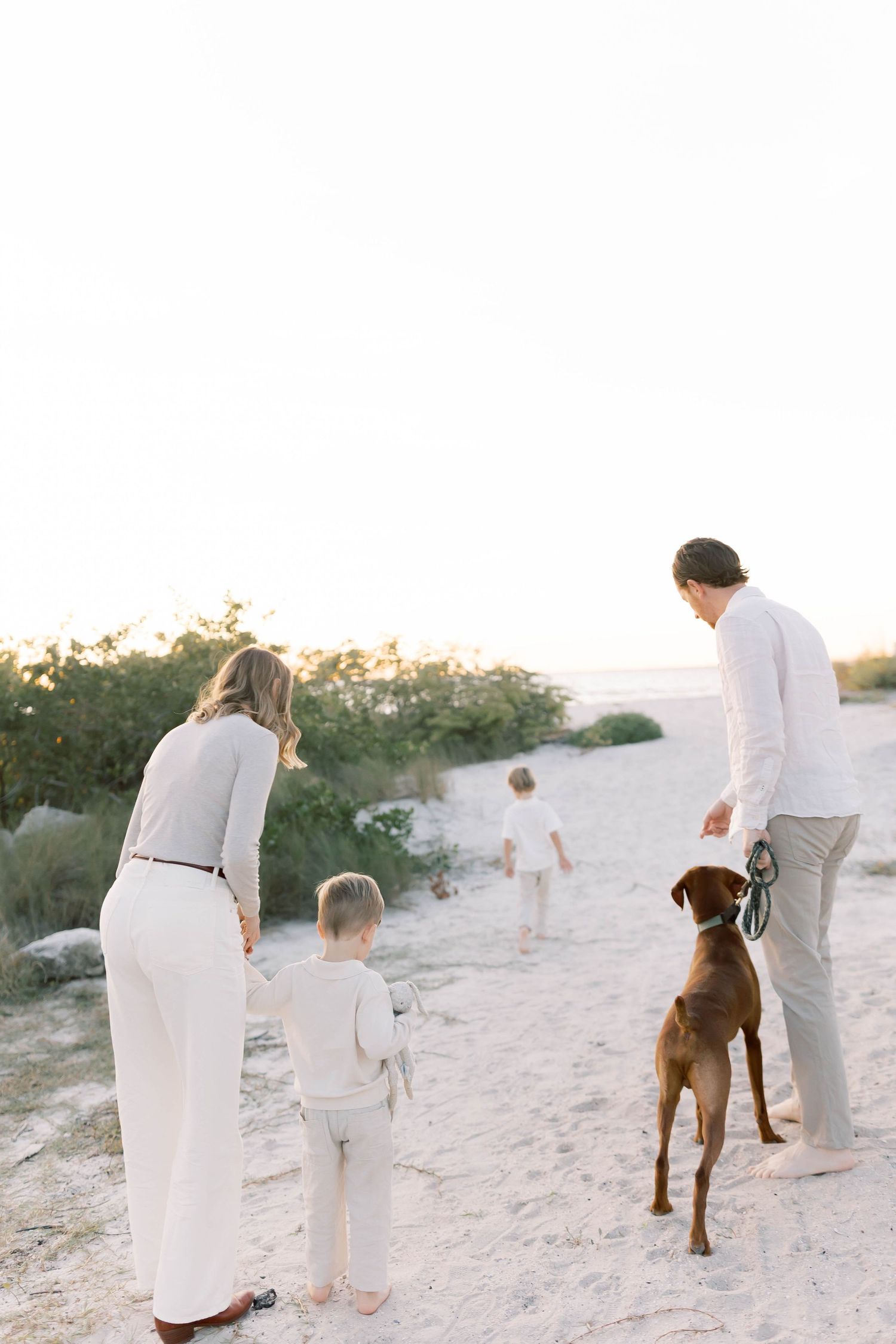 Family and brown dog explore beach together wearing coordinated white and beige outfits.