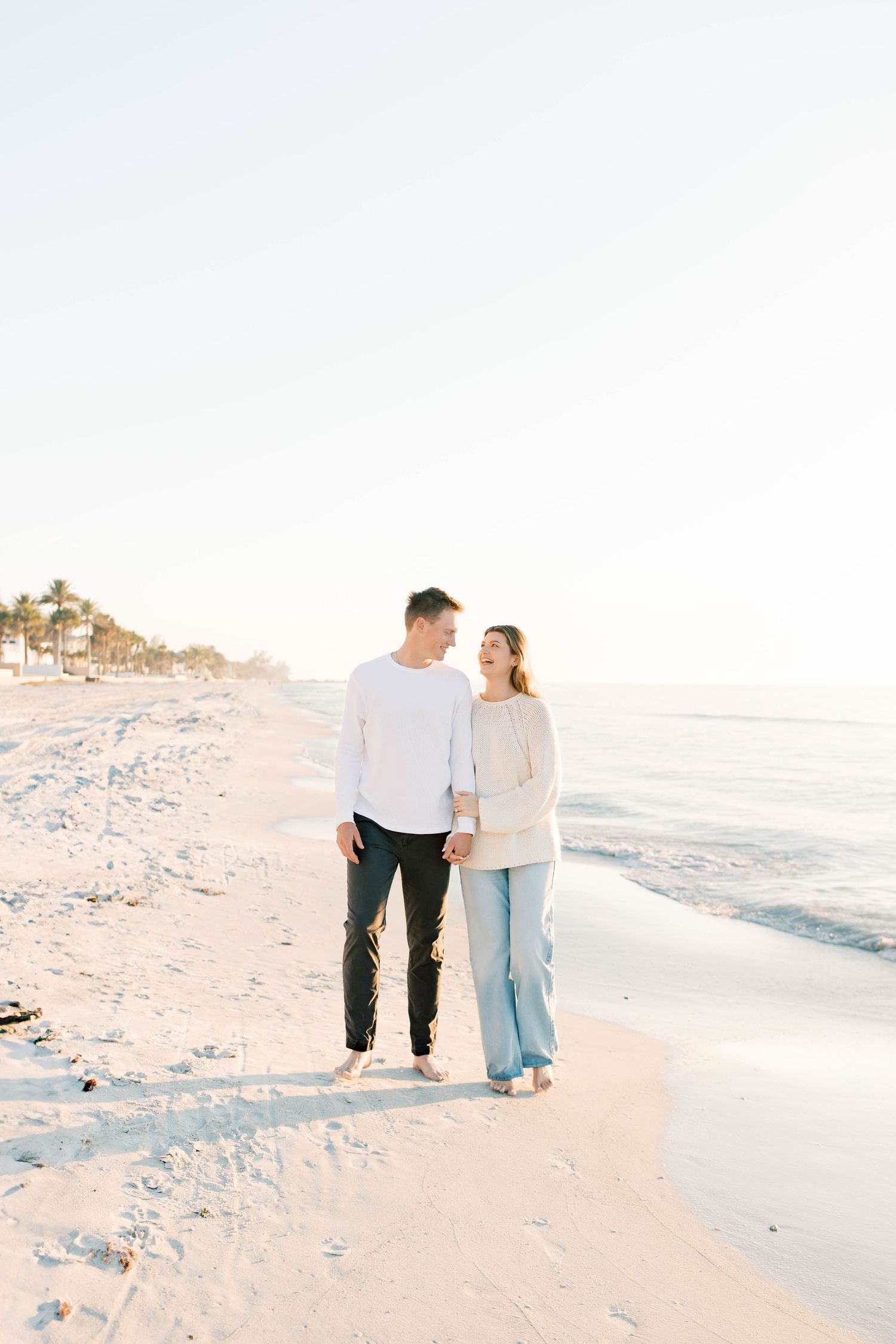 Two people walk together on a peaceful beach during sunset with coastal homes in the distance.