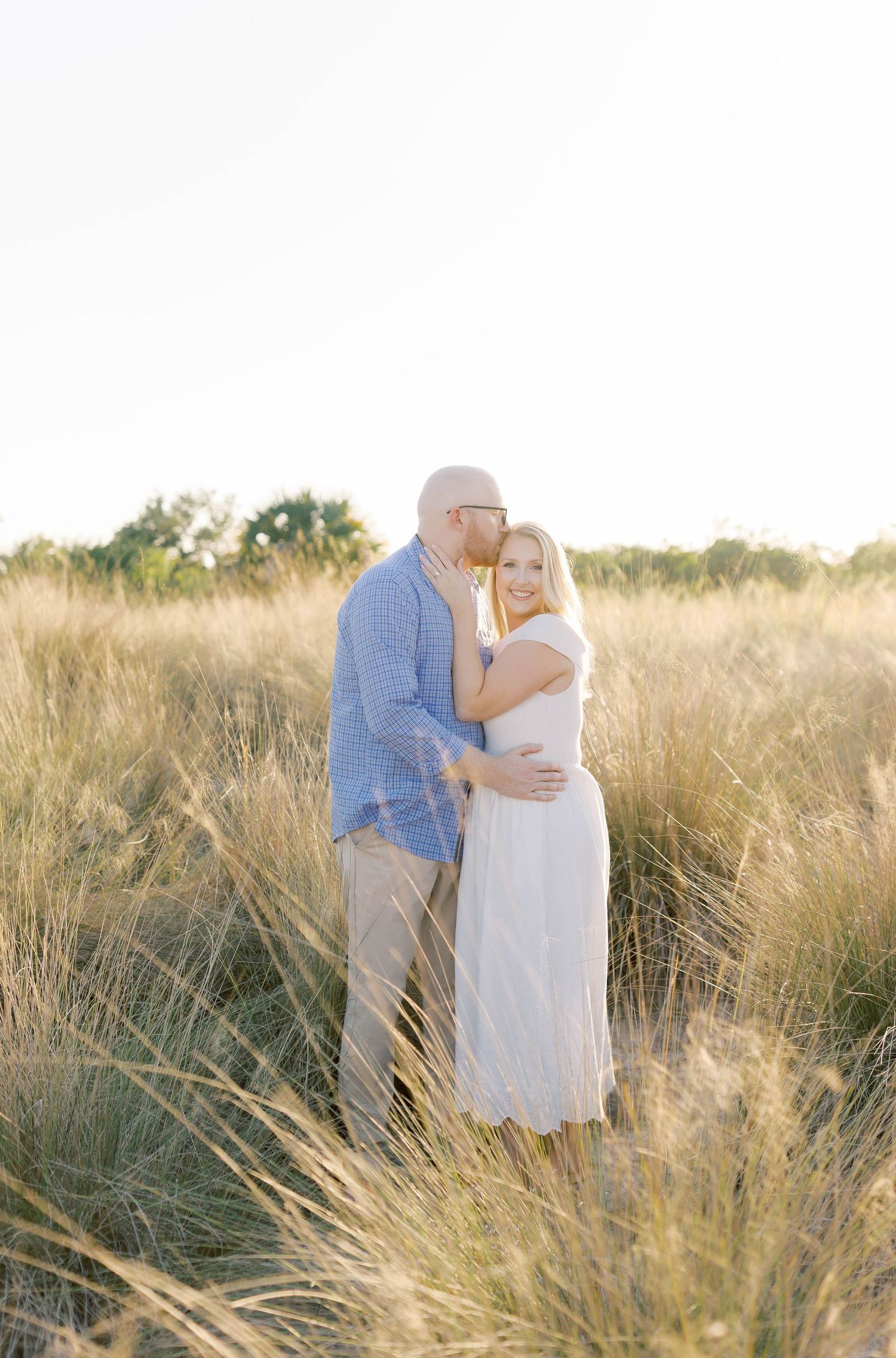 Couple embraces while standing among tall grass during a golden sunset photoshoot.