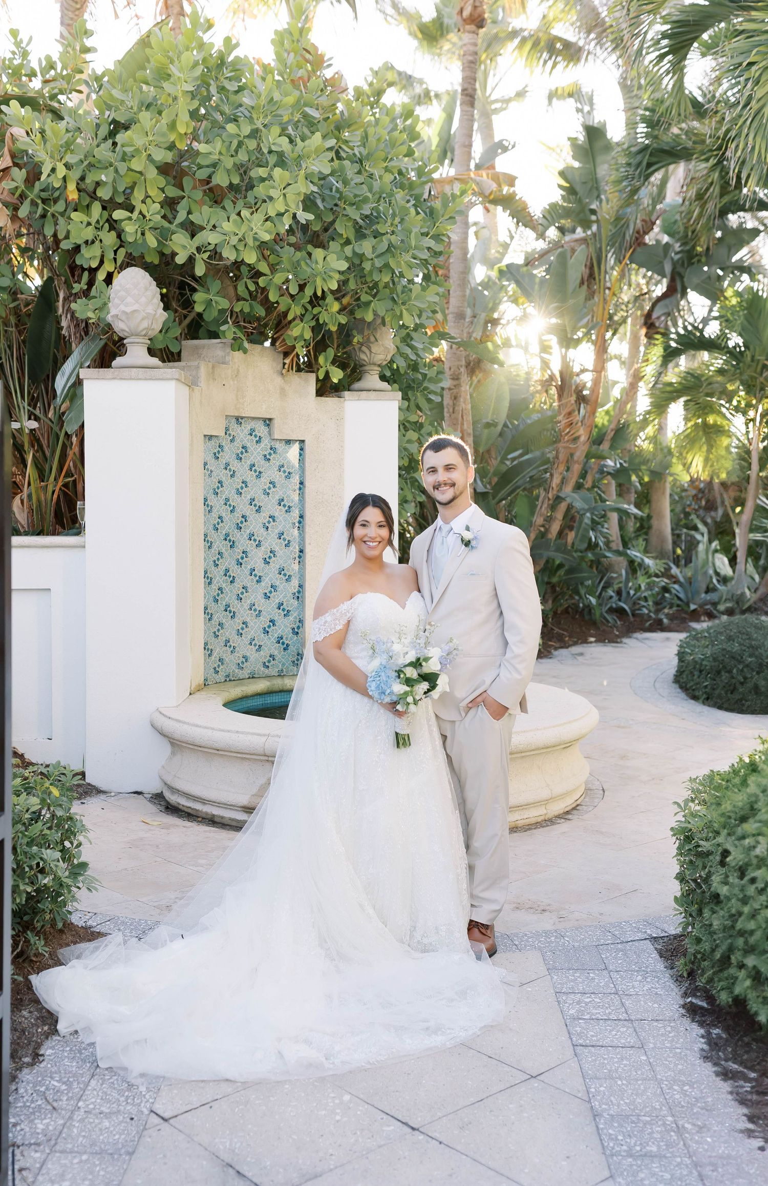 Smiling bride and groom pose together in a sunlit tropical garden beside an ornate tile fountain.