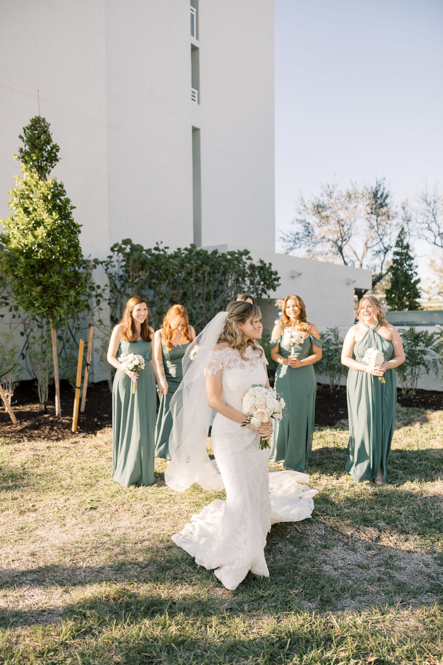 Wedding party in sage green dresses poses outdoors against a white wall during a sunny day.