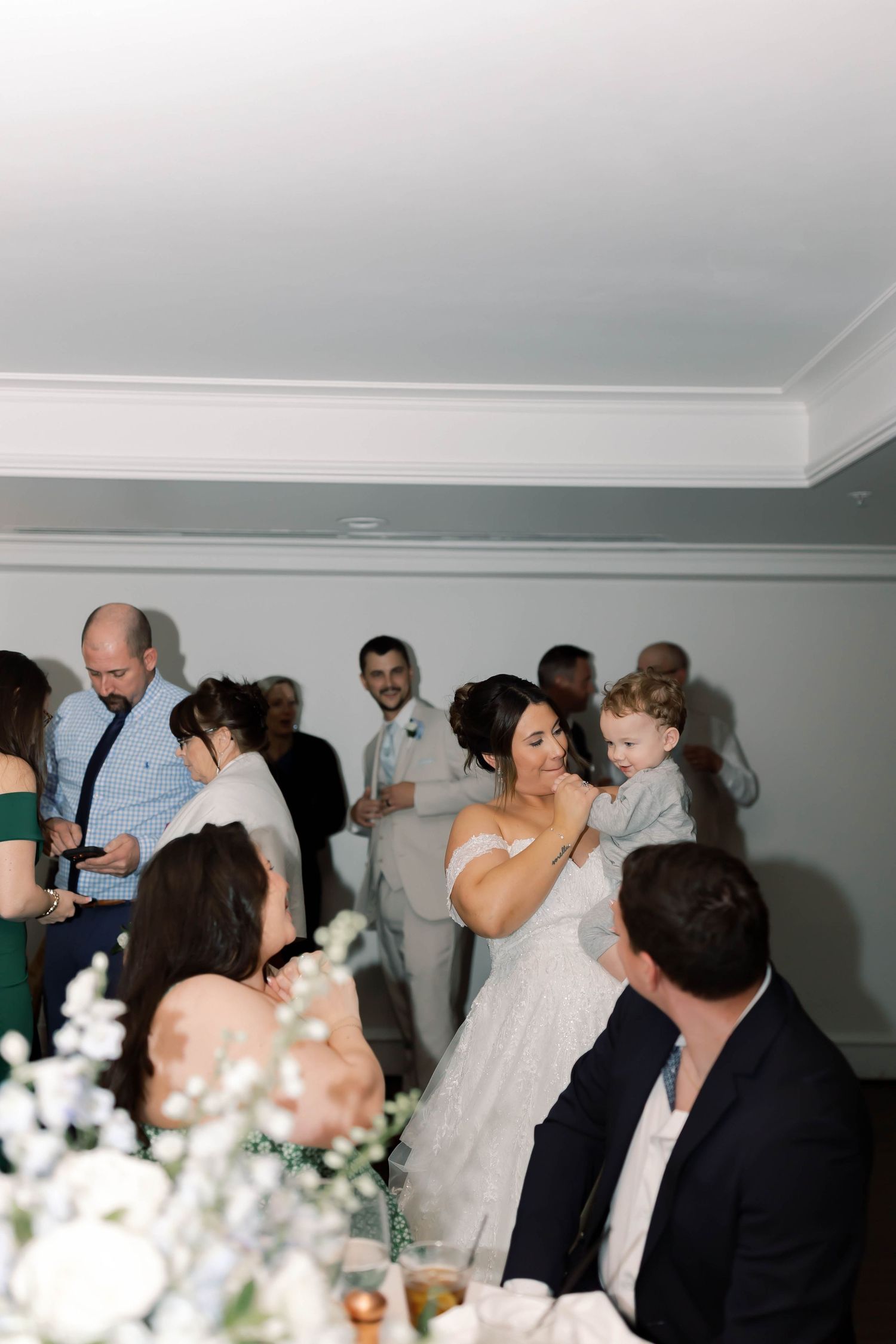 Bride in lace gown holds a young child while mingling with wedding guests during indoor reception celebration.